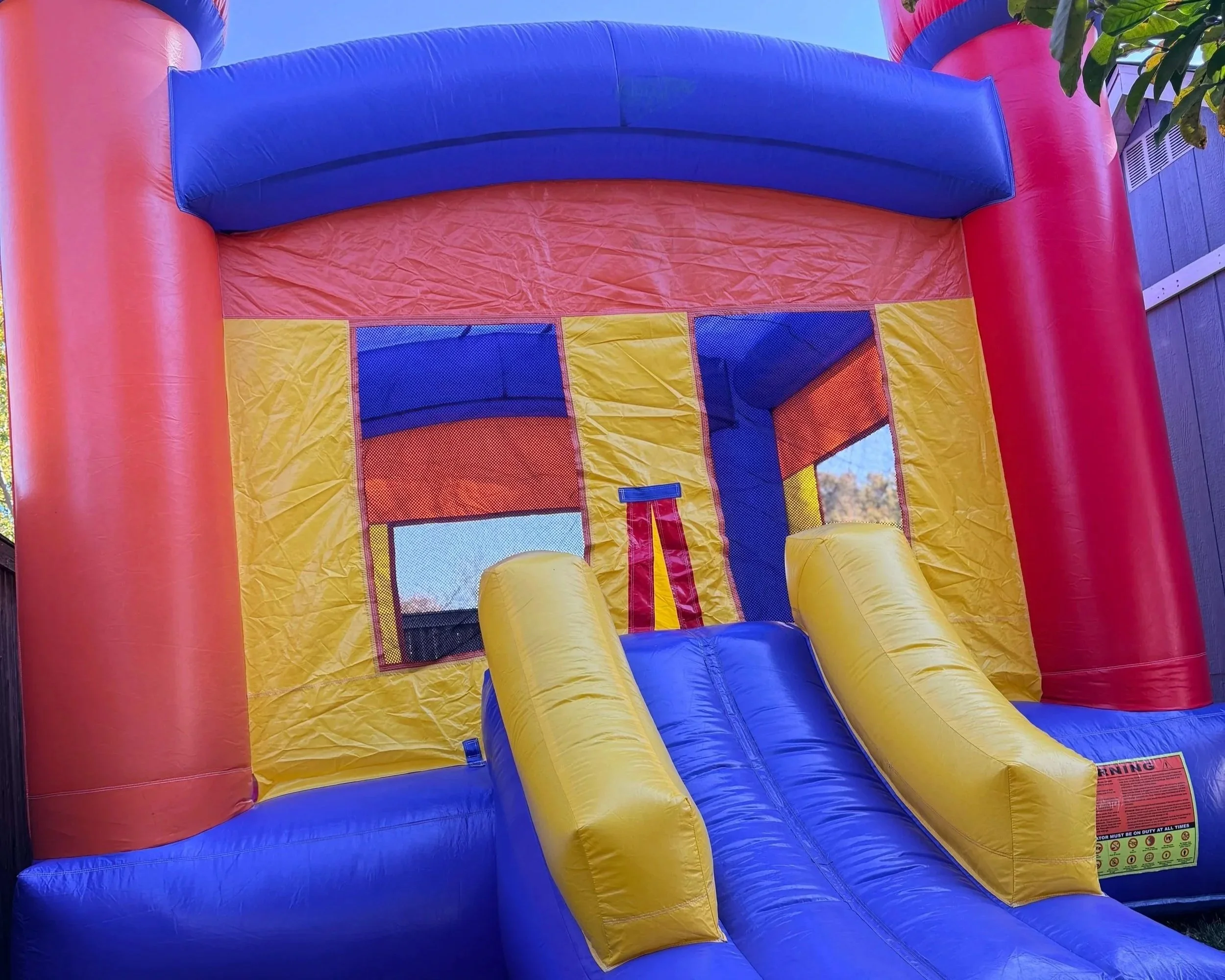 Colorful inflatable bounce house with two yellow slides, red, blue, and yellow panels, mesh windows, and a warning label at the bottom right corner.