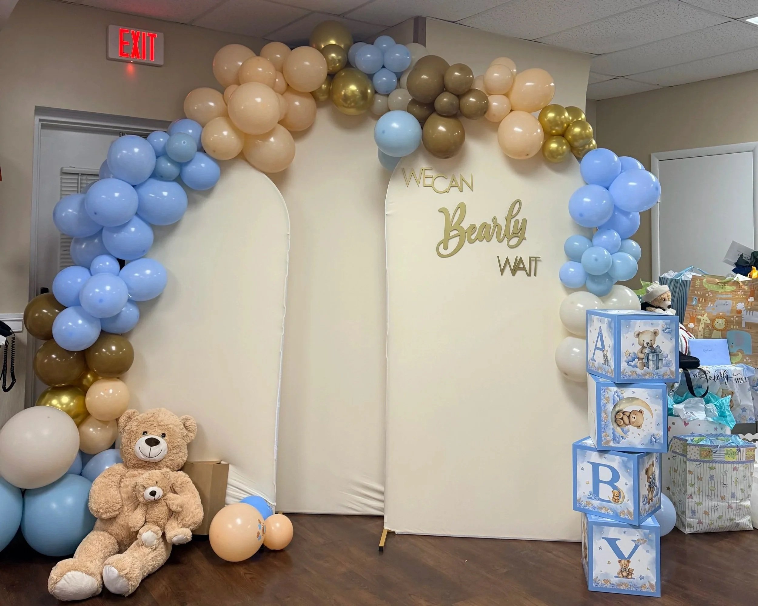 Baby shower decoration with a balloon arch in blue, beige, gold, and brown colors, teddy bears, and themed gift boxes with alphabet letters.