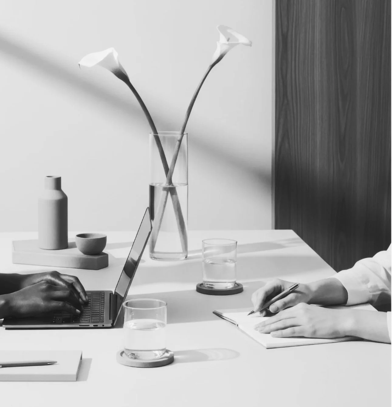 Two people working at a table, one typing on a laptop and the other writing in a notebook, with glasses of water and decorative vases on the table.
