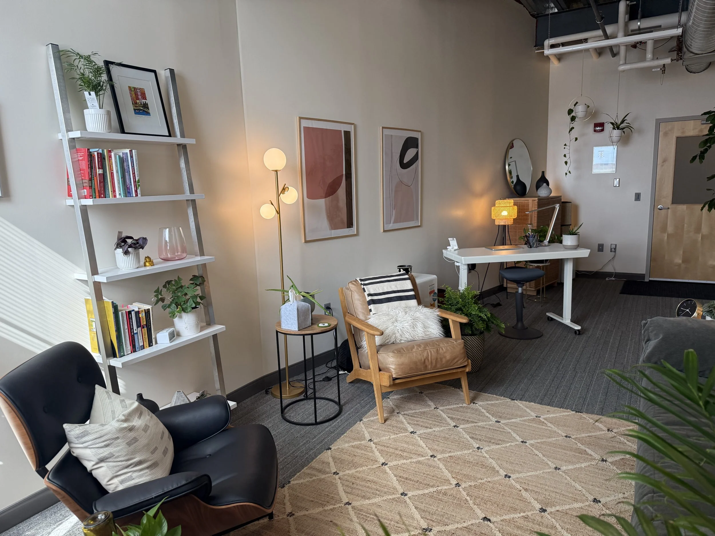 Modern office or living space with bookshelves, artwork, plants, a leather armchair, desk, and various decorative items.