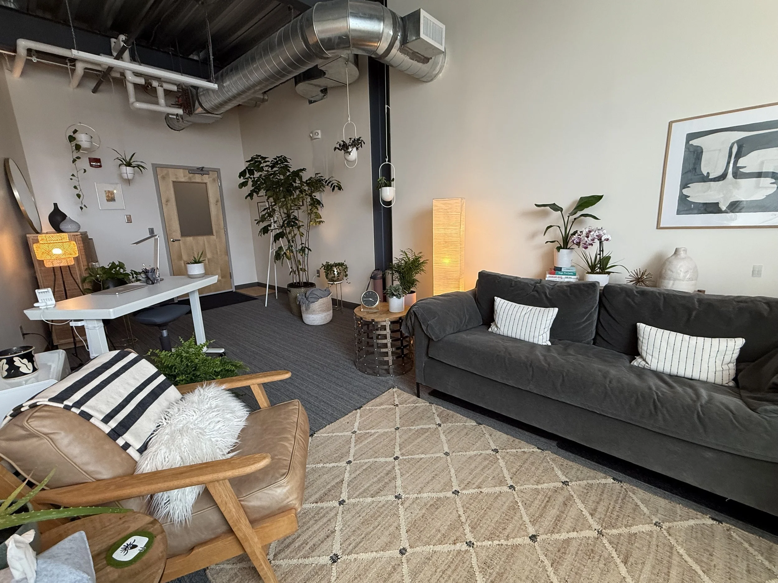 Cozy modern living room with dark gray sofa, beige patterned rug, and wooden armchair with striped pillow and fluffy white cushion; corner with tall plants, warm floor lamp, and black and white framed artwork on light-colored wall.