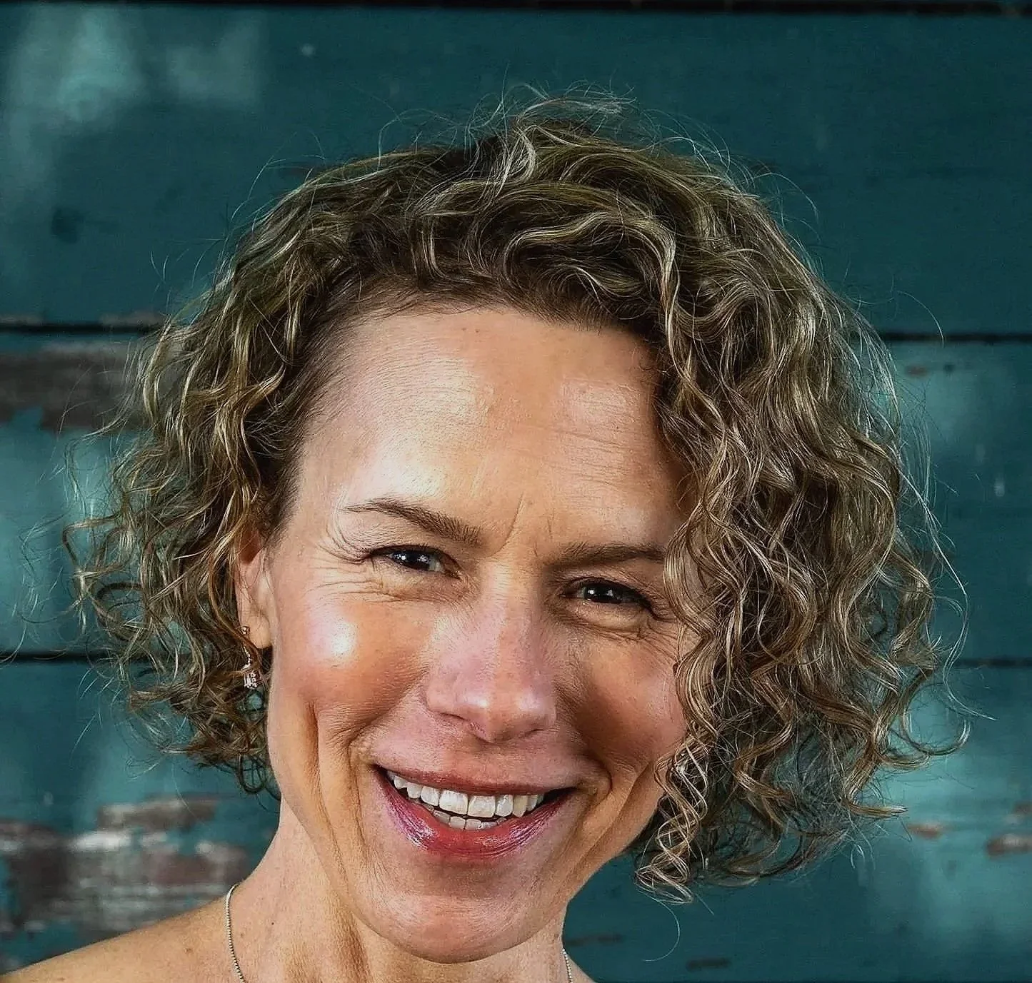 A smiling woman with curly short hair standing against a wood-paneled background.