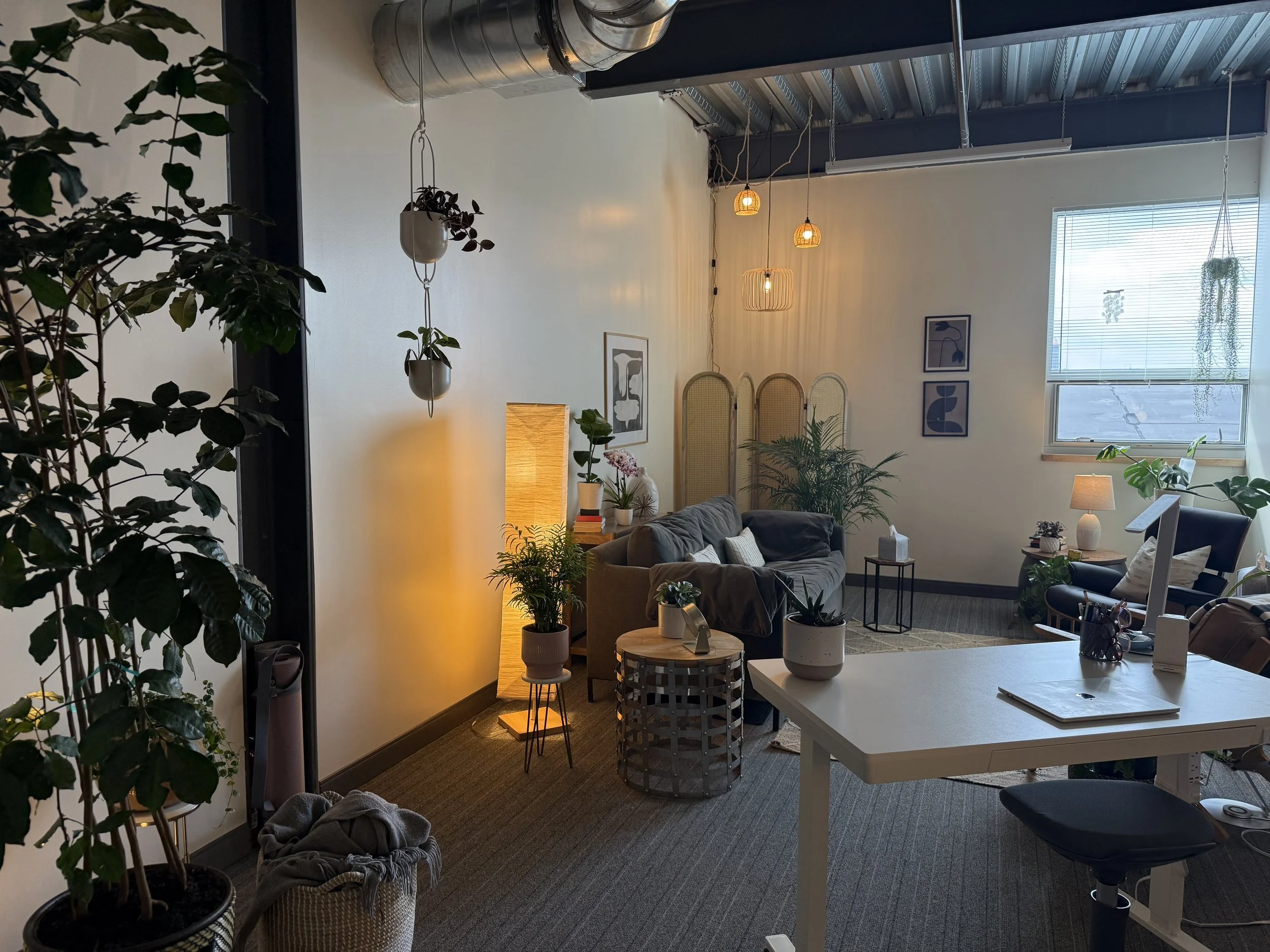 A cozy, well-lit living room with a beige sofa, armchair, and various plants. There is a white table with a notebook, a lamp, and plant pots. Warm lighting from lamps and hanging lights creates a comfortable atmosphere. A large window with blinds lets in natural light.