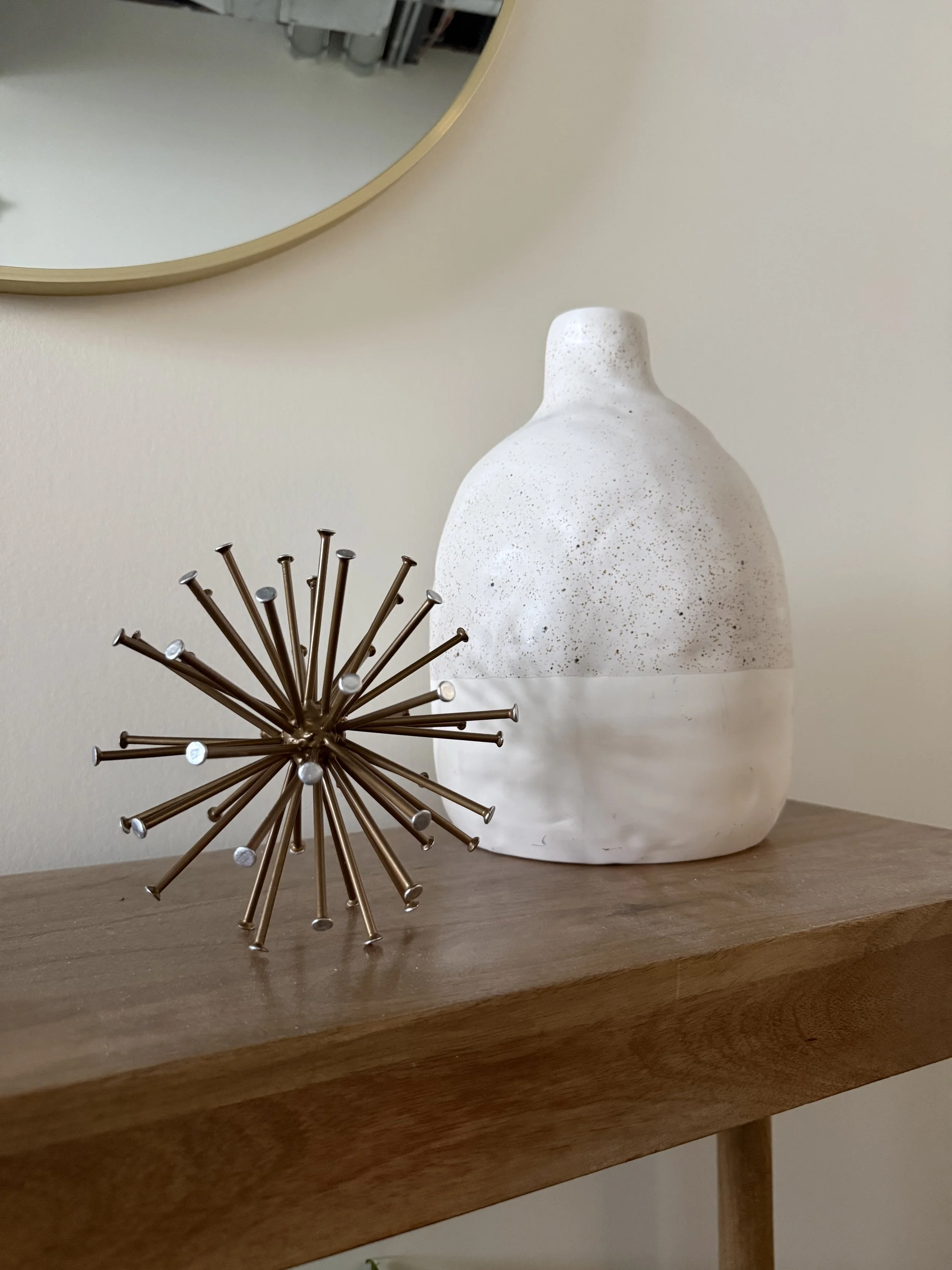 Decorative interior decor on a wooden shelf, including a white textured ceramic vase and a metallic spherical sculpture with multiple rods, with a mirror partially visible in the background.