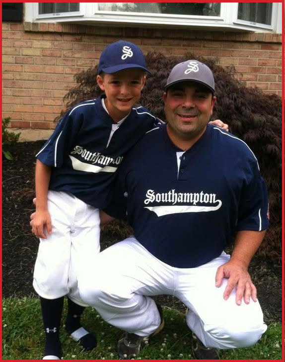 A man and a boy in baseball uniforms posing outdoors in front of a brick house. The boy is wearing a blue cap and a jersey, and the man is wearing a gray cap and matching jersey, both with the word "Southampton". The boy is leaning on the man's shoulder, and they are smiling.