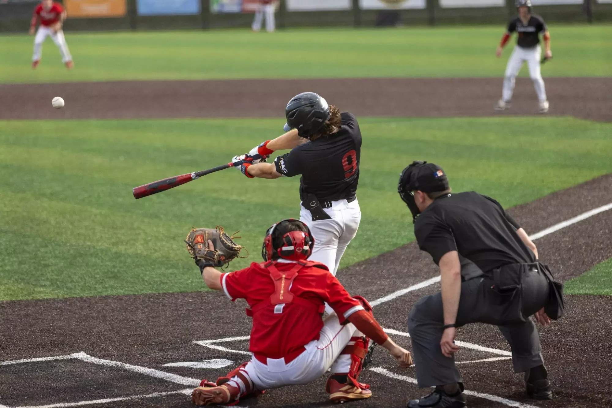Baseball batter in black jersey swings at a pitch as the catcher in red gear lunges to catch the ball near home plate, with an umpire monitoring the play, on a baseball field.