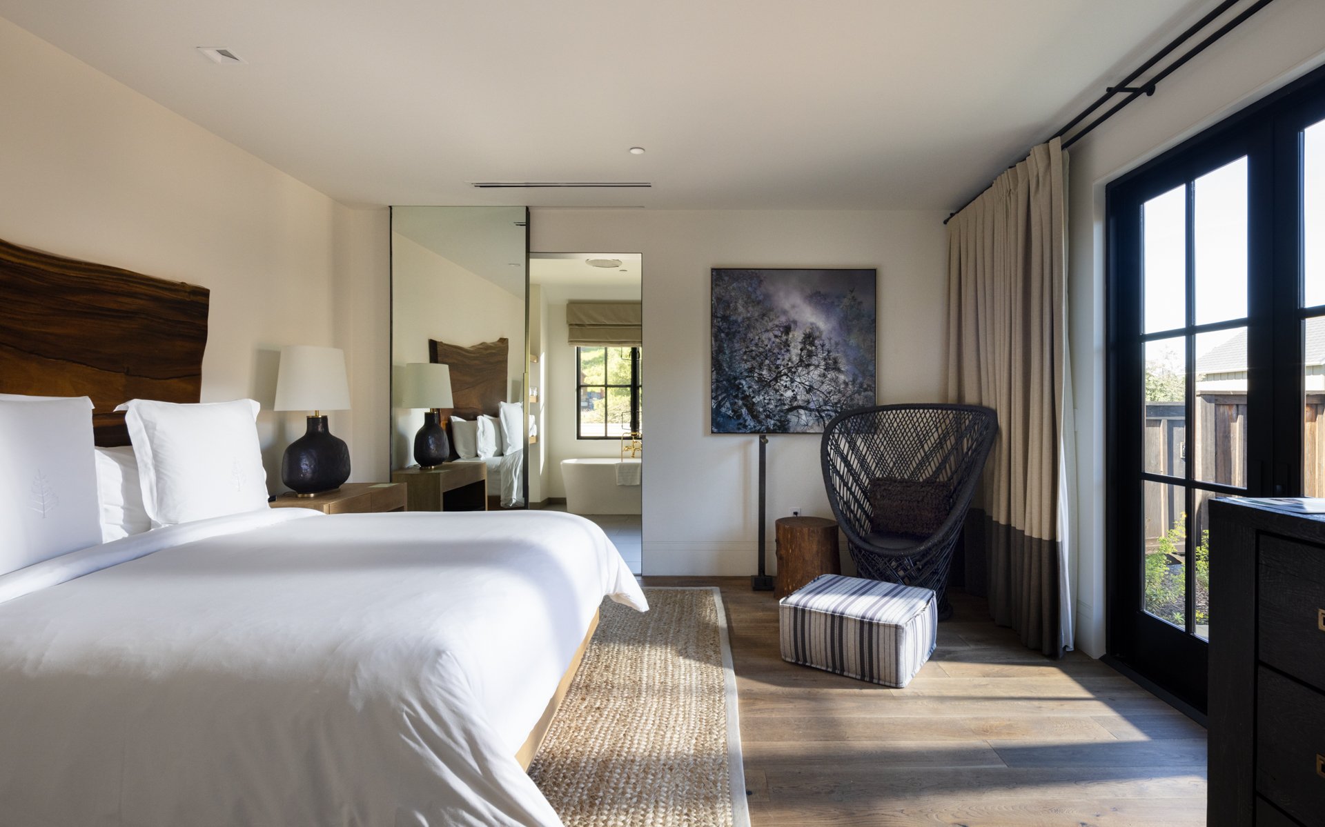 Hotel room with a large bed, nightstands with lamps, a decorative chair, and a sliding glass door leading outside, with a view of a fenced yard.