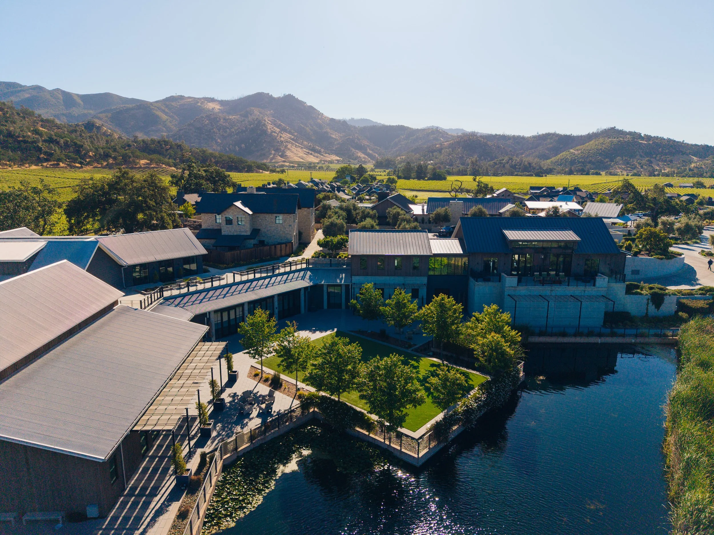Aerial view of a modern residential community with a central water feature, surrounded by lush green trees and mountains in the background, under a clear blue sky.