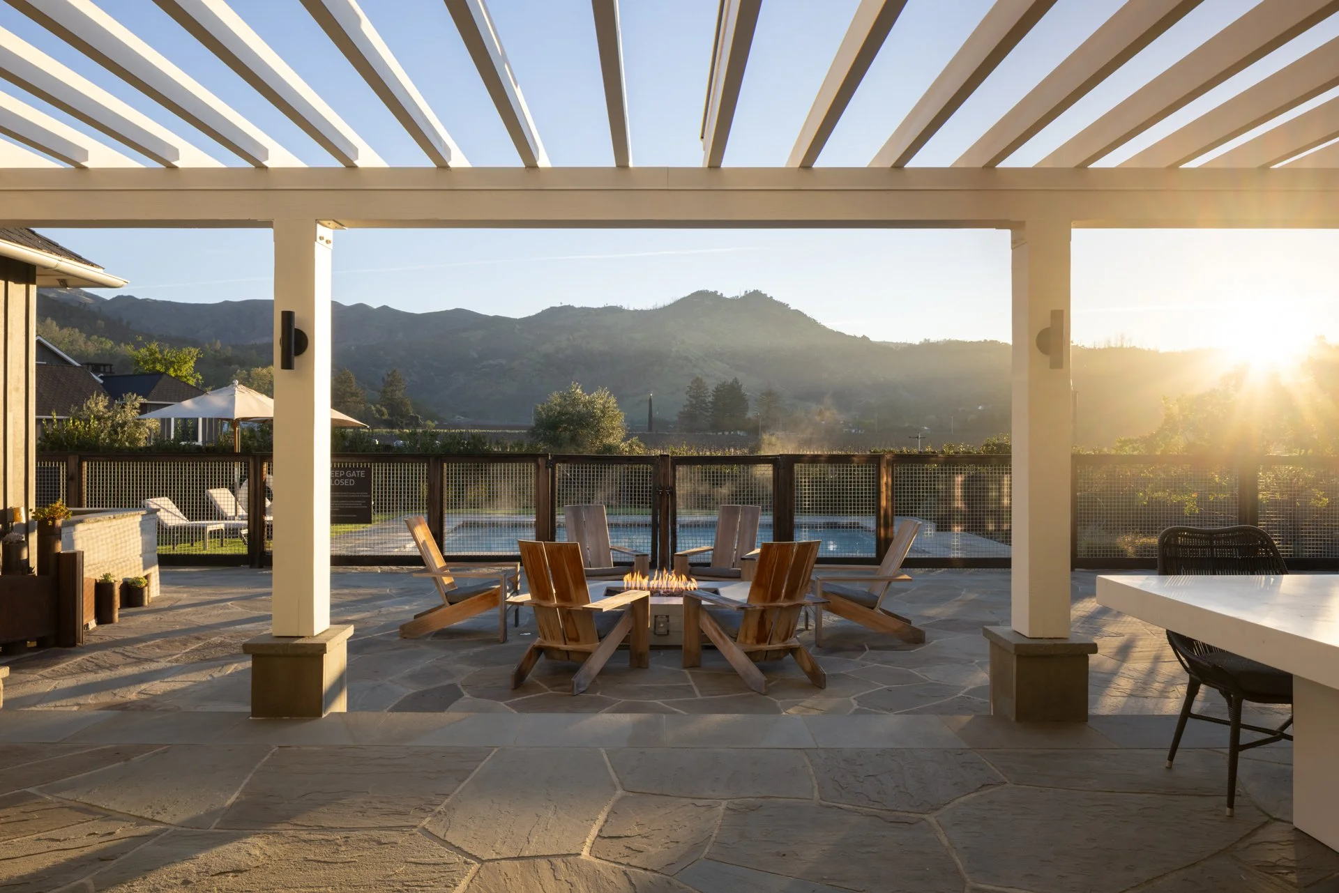 An outdoor patio area with wooden chairs arranged around a fire pit, overlooking a pool, with mountains in the background and sunlight shining from the right.