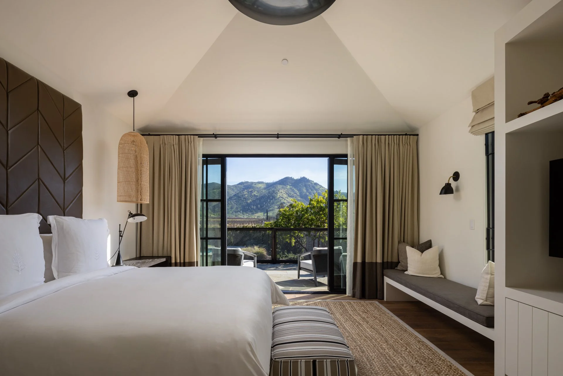 A bedroom with a view of mountains outside, featuring a large bed, beige curtains, patio furniture, and a balcony.