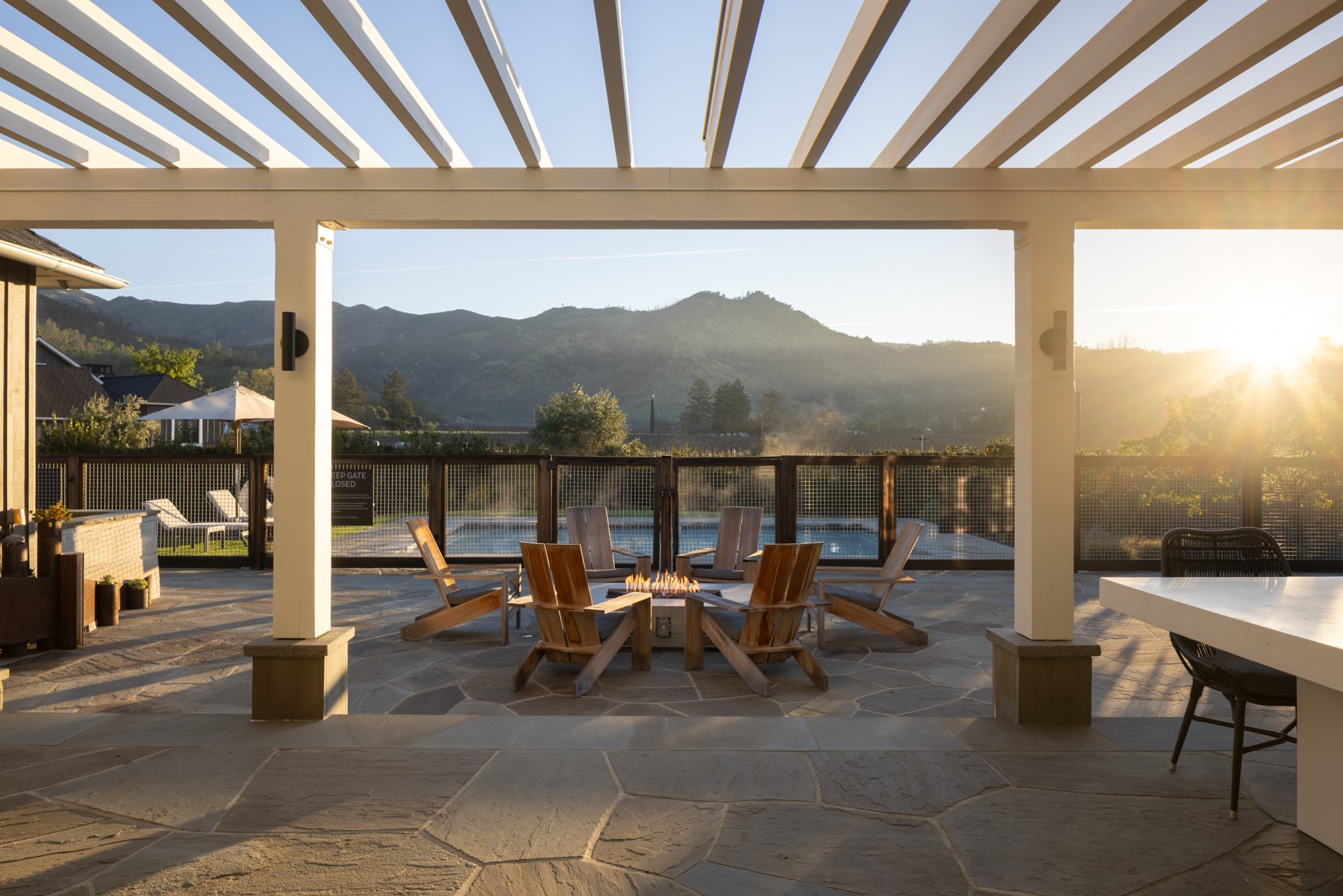 Sunset view from a patio with a fire pit surrounded by six wooden chairs, overlooking a fenced pool and green mountains in the background.