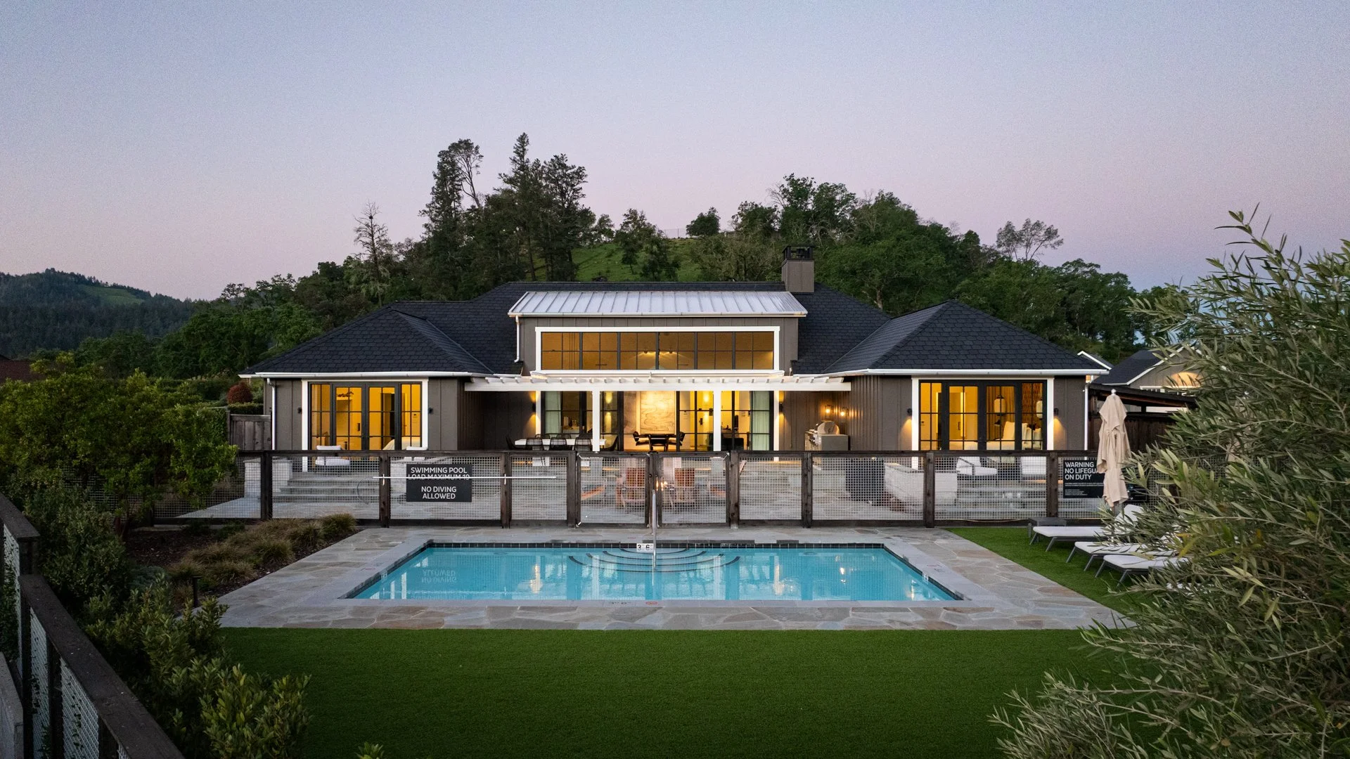 Welcoming house with large glass windows, illuminated interior, backyard swimming pool, surrounded by lush green landscape and hills at dusk.