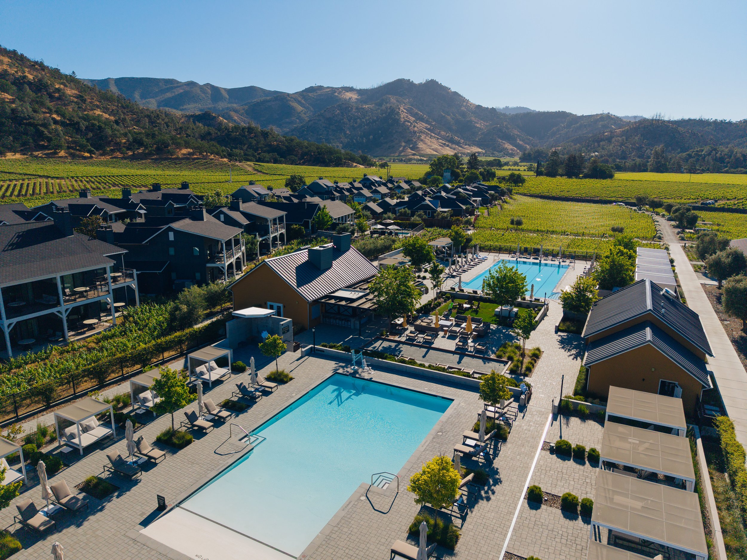 Aerial view of a modern residential complex with two swimming pools, surrounded by lounge chairs, umbrellas, and trees, set against rolling hills and vineyards.
