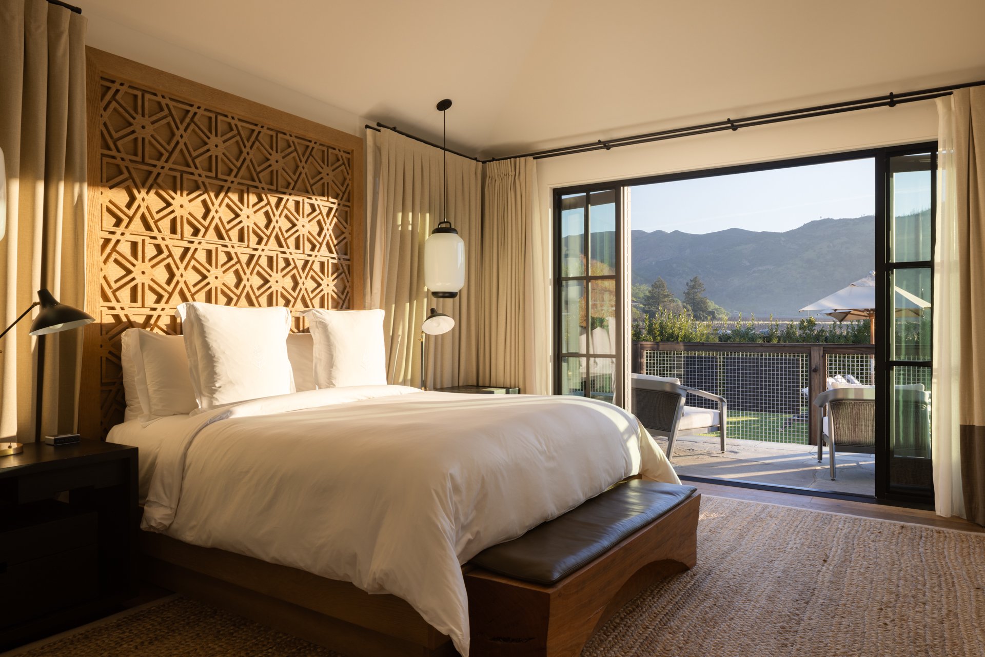 A bedroom with a large bed dressed with white linens and pillows, a wooden headboard with decorative lattice design, a pendant light hanging from the ceiling, and sliding glass doors that open to a balcony with outdoor furniture, overlooking a mounta