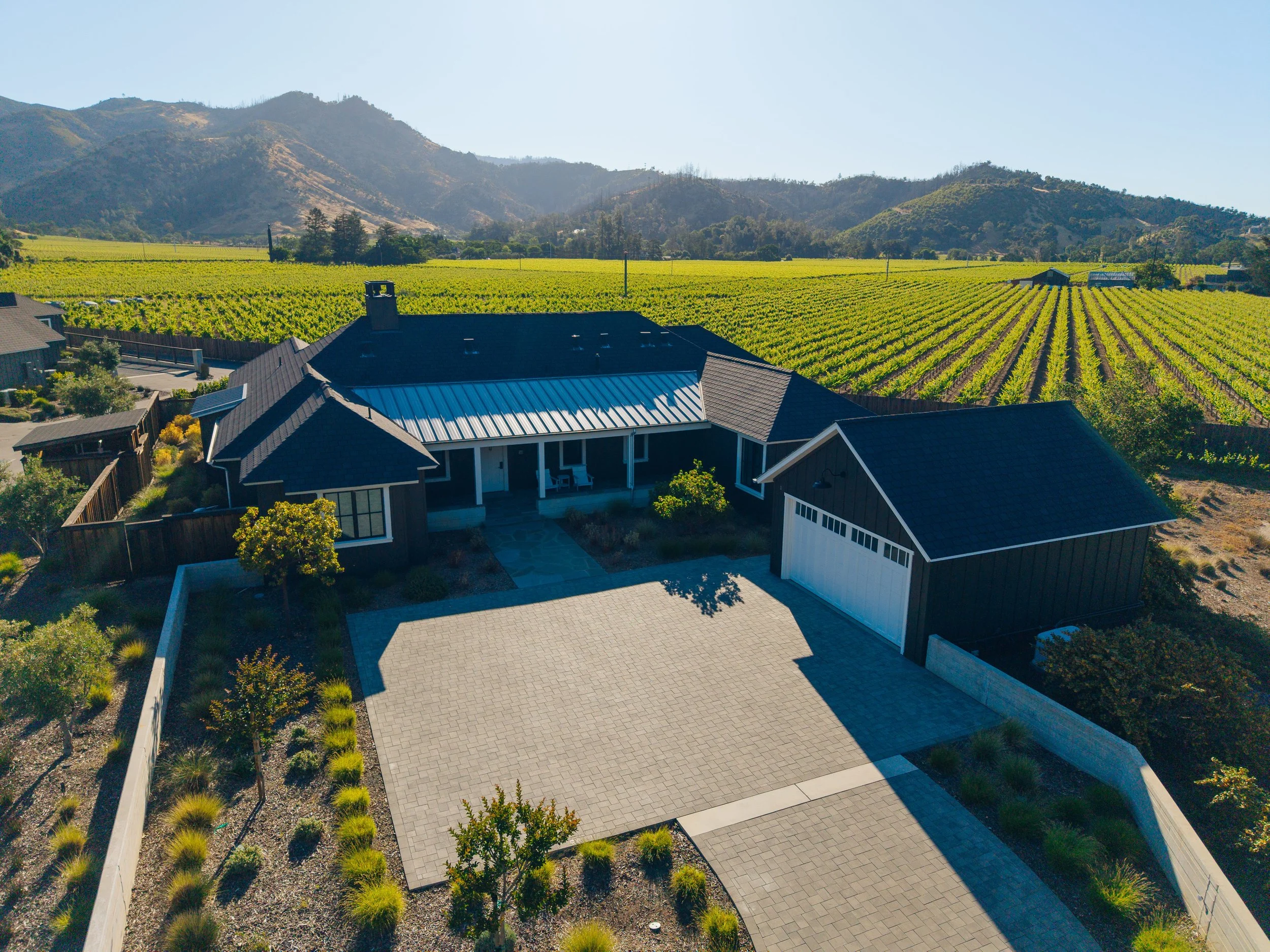 Aerial view of a modern house with a large driveway, surrounded by a landscaped yard with small trees and bushes, with extensive vineyards and rolling hills in the background during daytime.