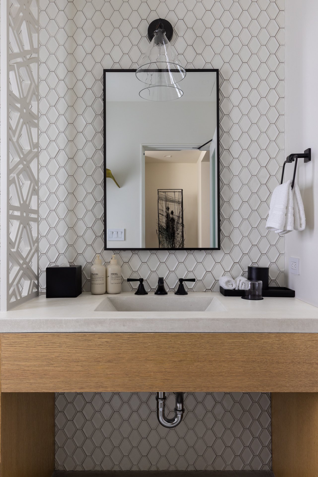 Modern bathroom vanity with a large rectangular mirror, hexagonal tile wall, black fixtures, white towels, black accessories, and visible plumbing beneath the sink.