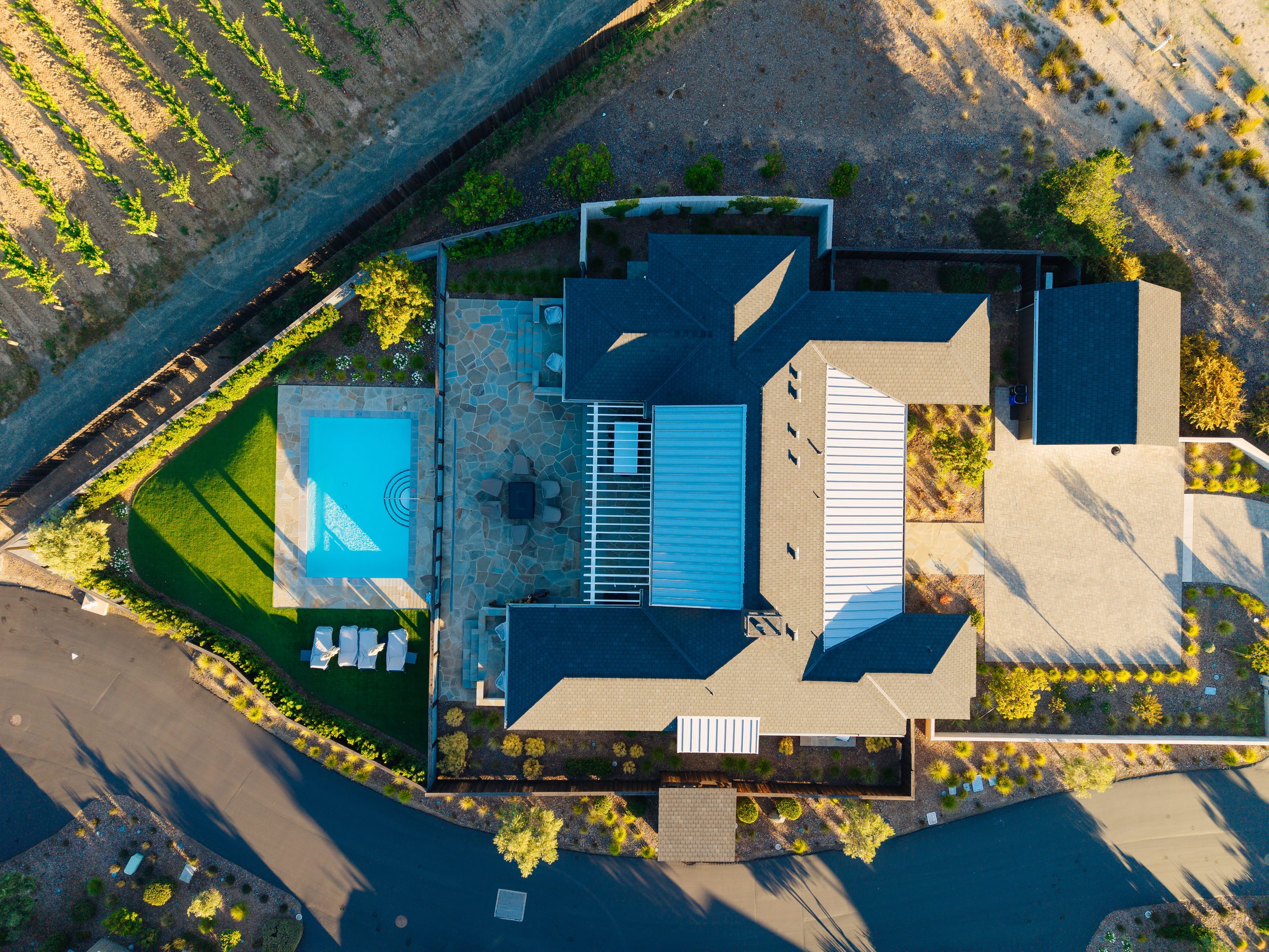 Aerial view of a house with a backyard featuring a swimming pool, patio furniture, and a landscaped yard, surrounded by a fence and a gravel driveway.