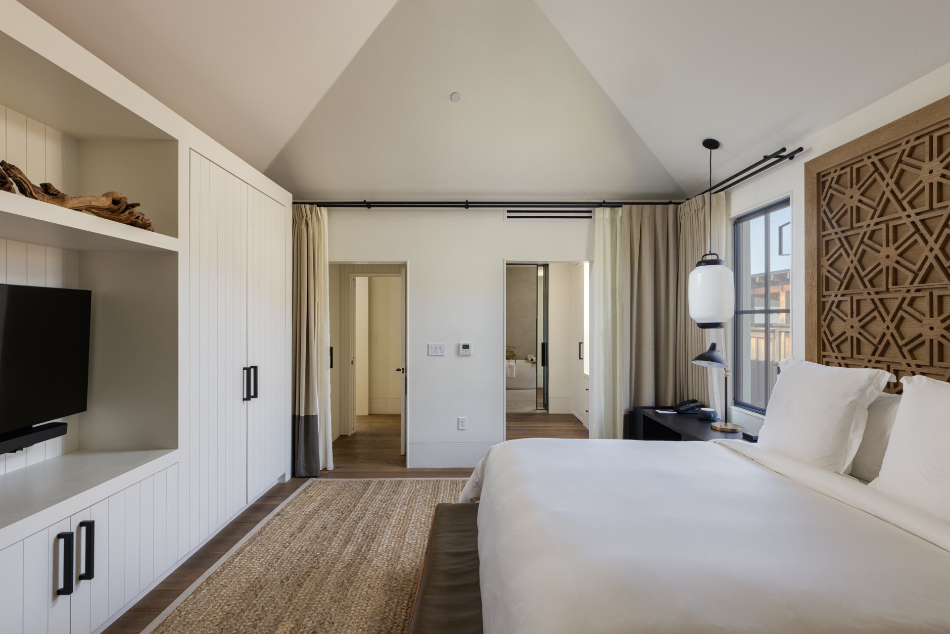 A modern hotel bedroom with a large bed, white bedding, a wooden headboard, a bedside table with a lamp, a wall-mounted television, beige curtains, and sliding glass doors leading to a balcony.