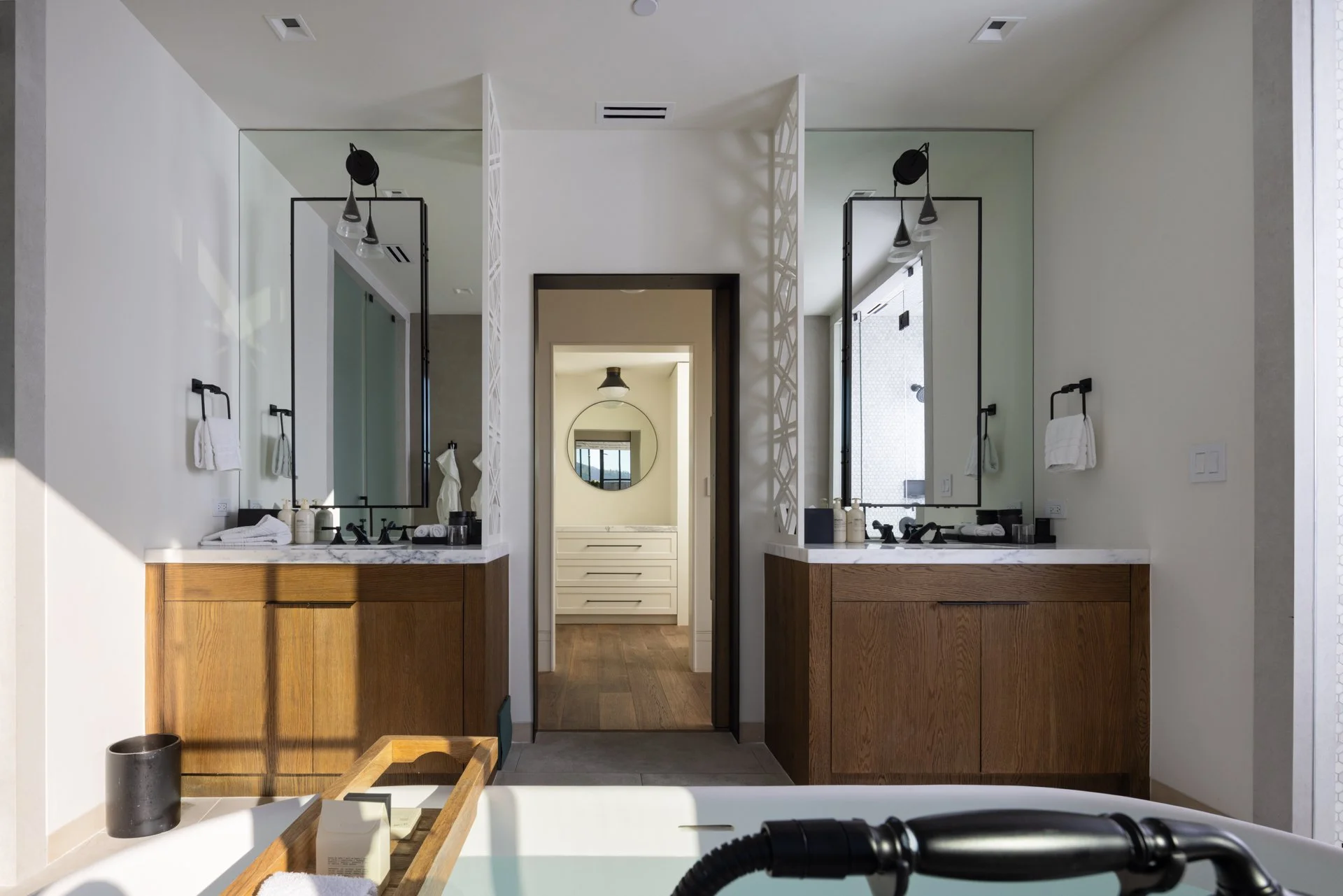 Modern bathroom with double vanity sinks, large mirrors, and wooden cabinetry, seen from the bathtub. There is a door leading to a walk-in closet or laundry area in the background.