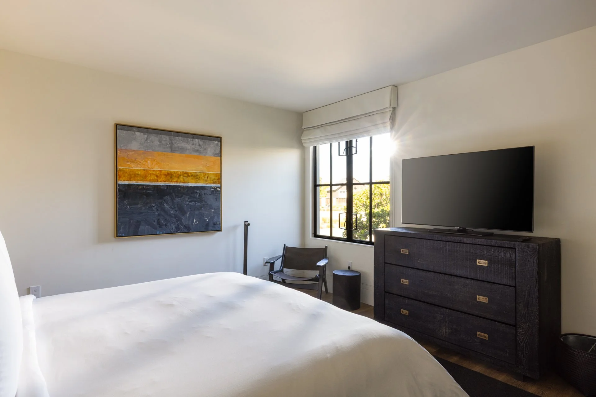 Bedroom with large bed, abstract wall art, window with sunlight, black dresser with TV, black chair, small black side table, white walls.