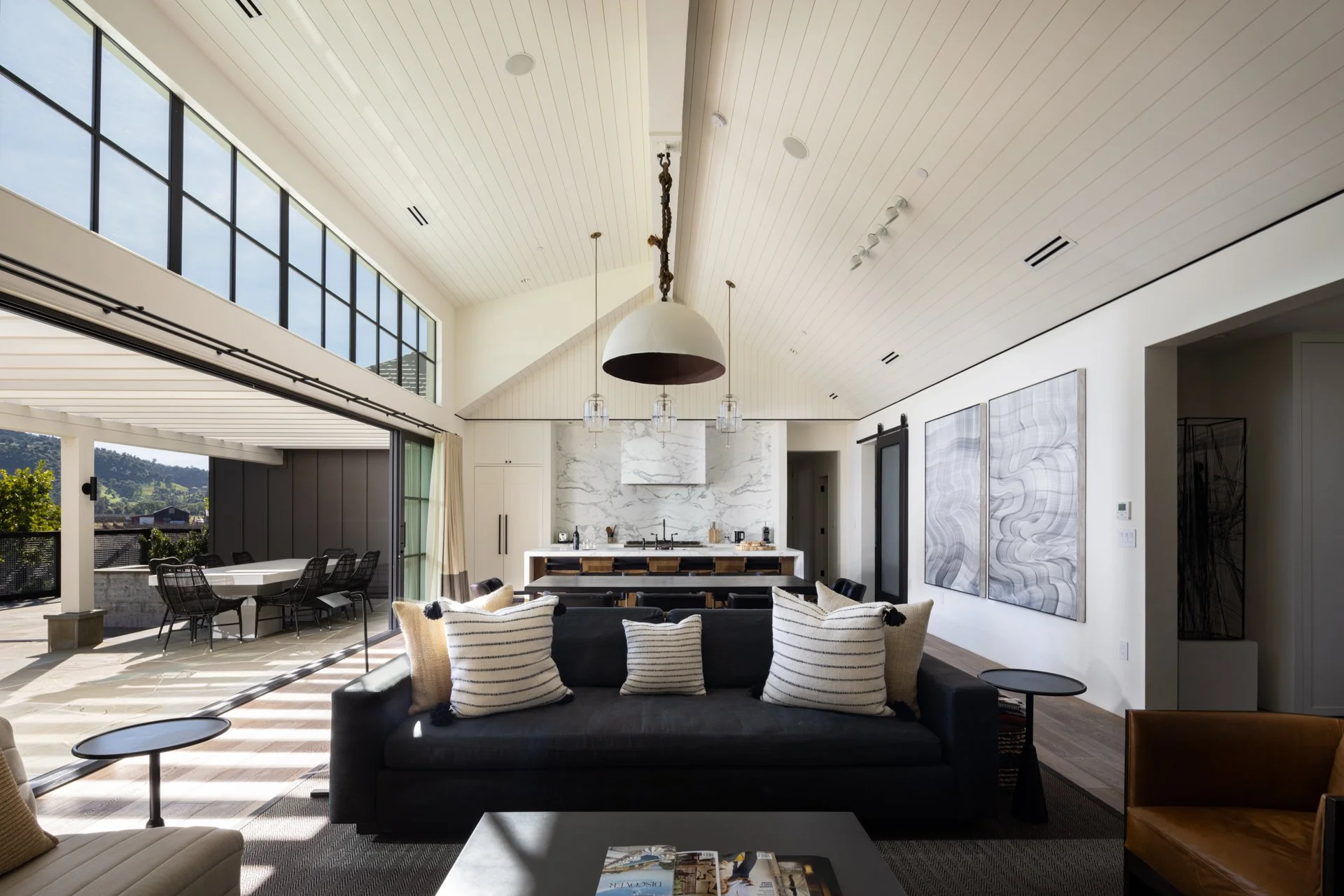 Open-concept living room with high white vaulted ceiling, large windows, modern art on white walls, black sofa with striped pillows, and views of outdoor patio with dining table.