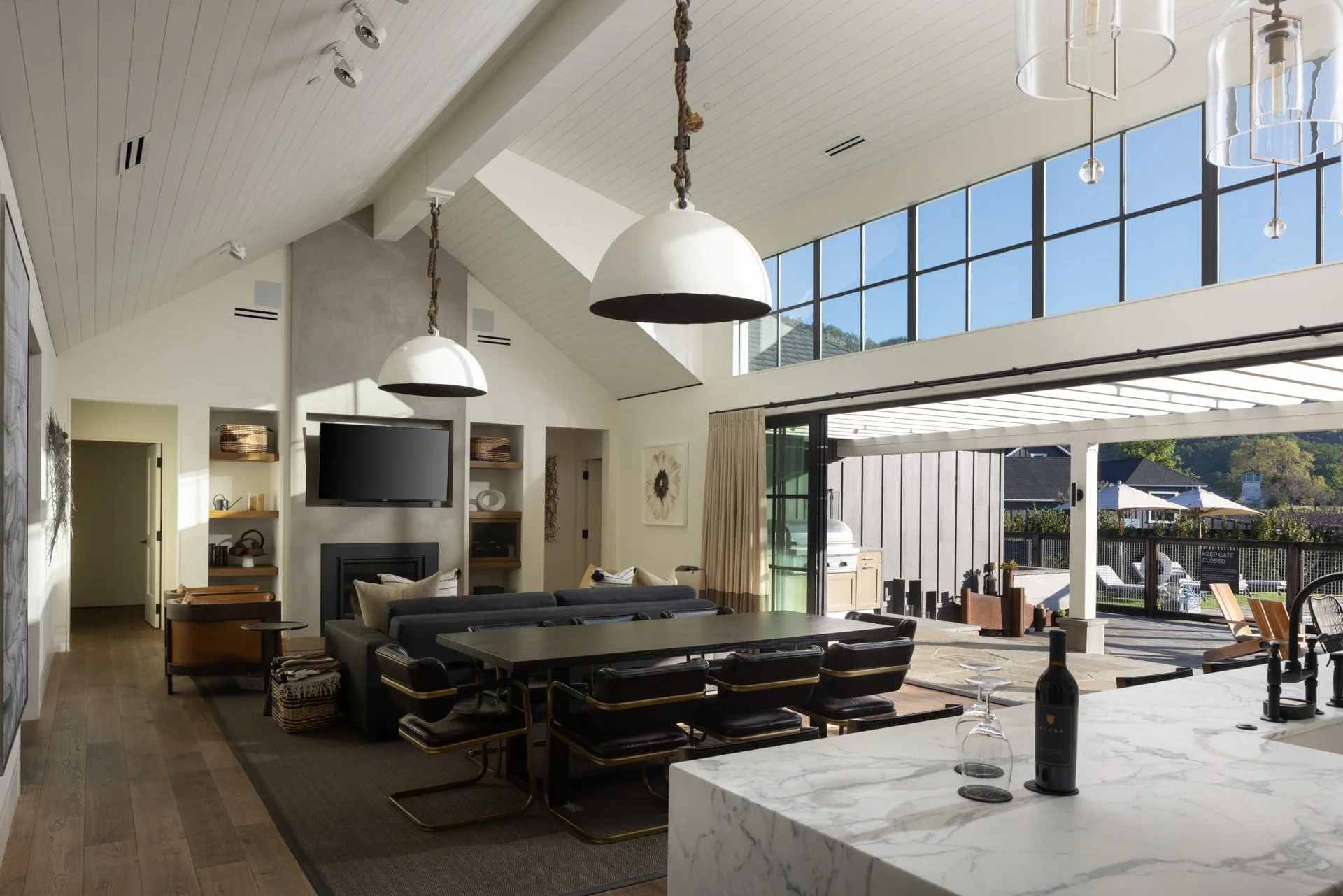 Open-concept living room with high vaulted ceiling, large windows, modern black and white furniture, a fireplace, and a view of a patio area outside.