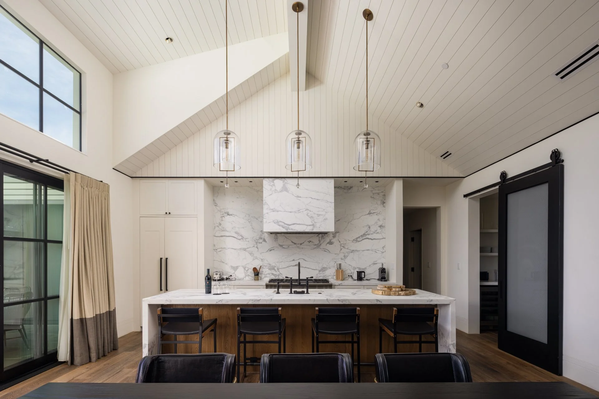 Modern kitchen with white marble island, black bar stools, and a marble backsplash. Large windows and beige curtains on the left, wood flooring, and ceiling lights hanging above the island.