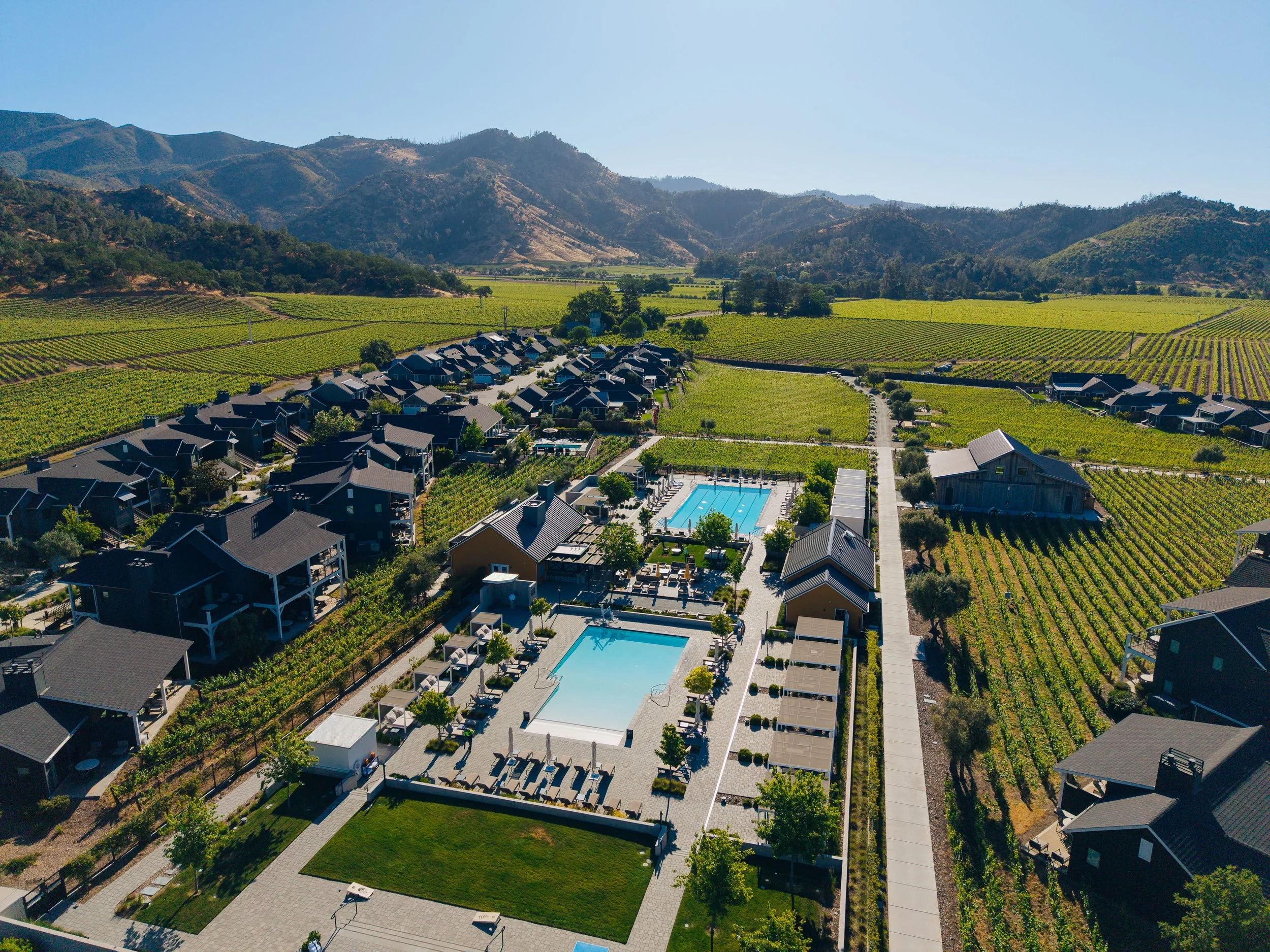 Aerial view of a community with houses, two swimming pools, and surrounding vineyards and mountains.