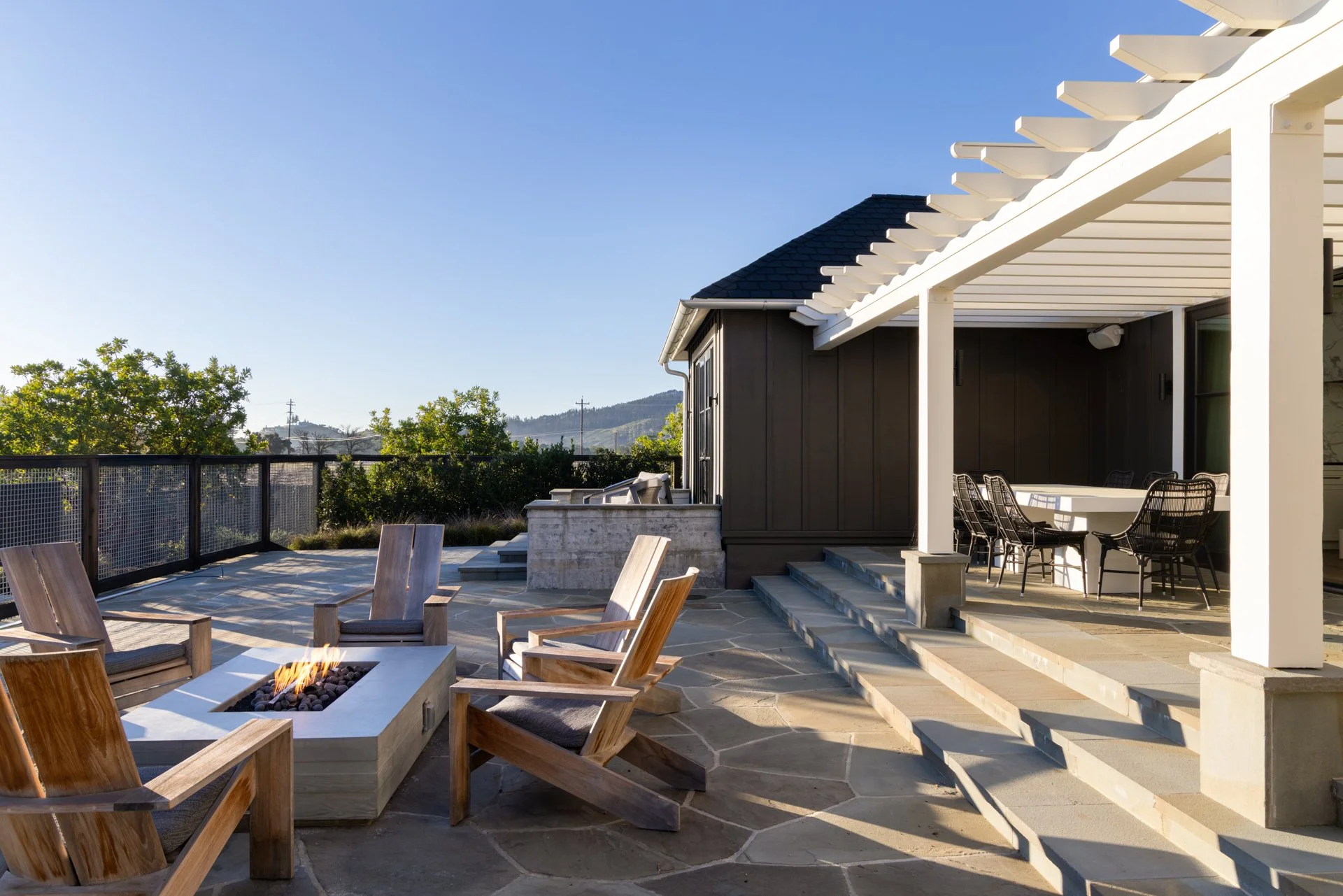 Outdoor patio with wooden chairs around a fire pit, adjacent to a modern house with a covered dining area and scenic landscape in the background.