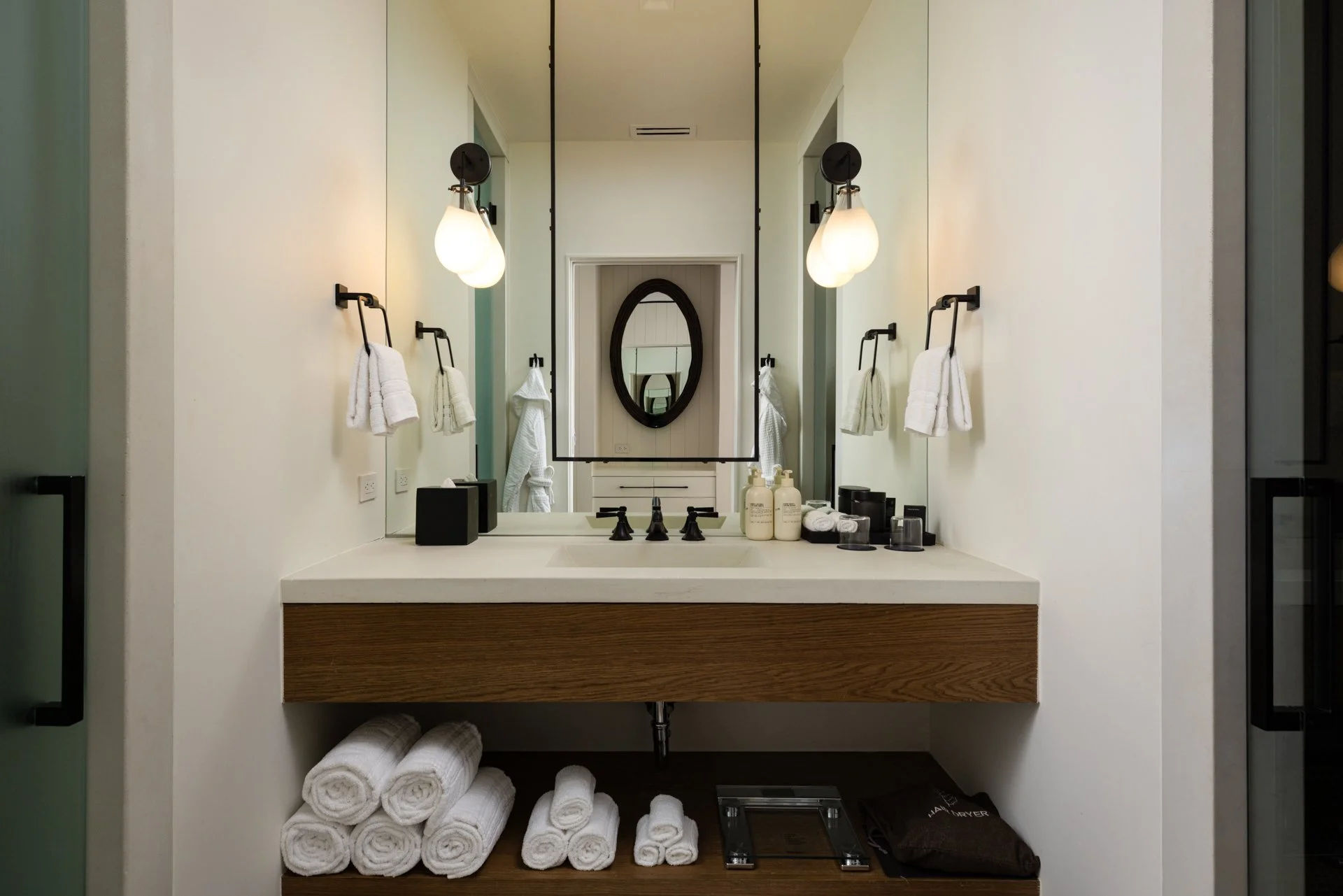 A modern bathroom vanity with a large mirror, surrounded by two wall-mounted light fixtures, white towels on black rings, and toiletries including soap bottles and jars. There is a shelf with rolled towels and a scale underneath.
