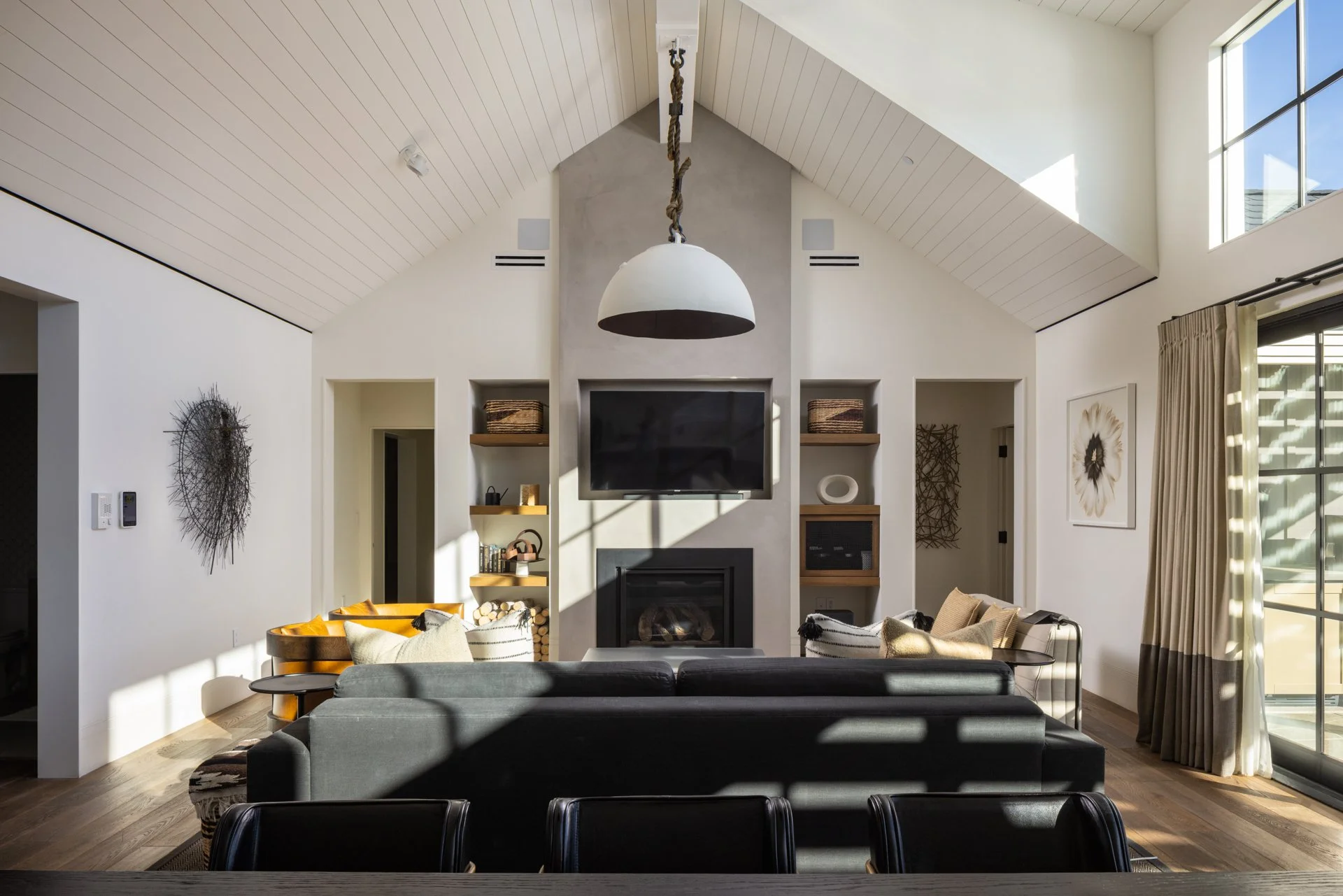 Living room with a high vaulted ceiling, a fireplace, a large flat-screen TV, and seating including a black sofa and patterned chairs. Large windows with curtains let in sunlight, and artwork decorates the walls.