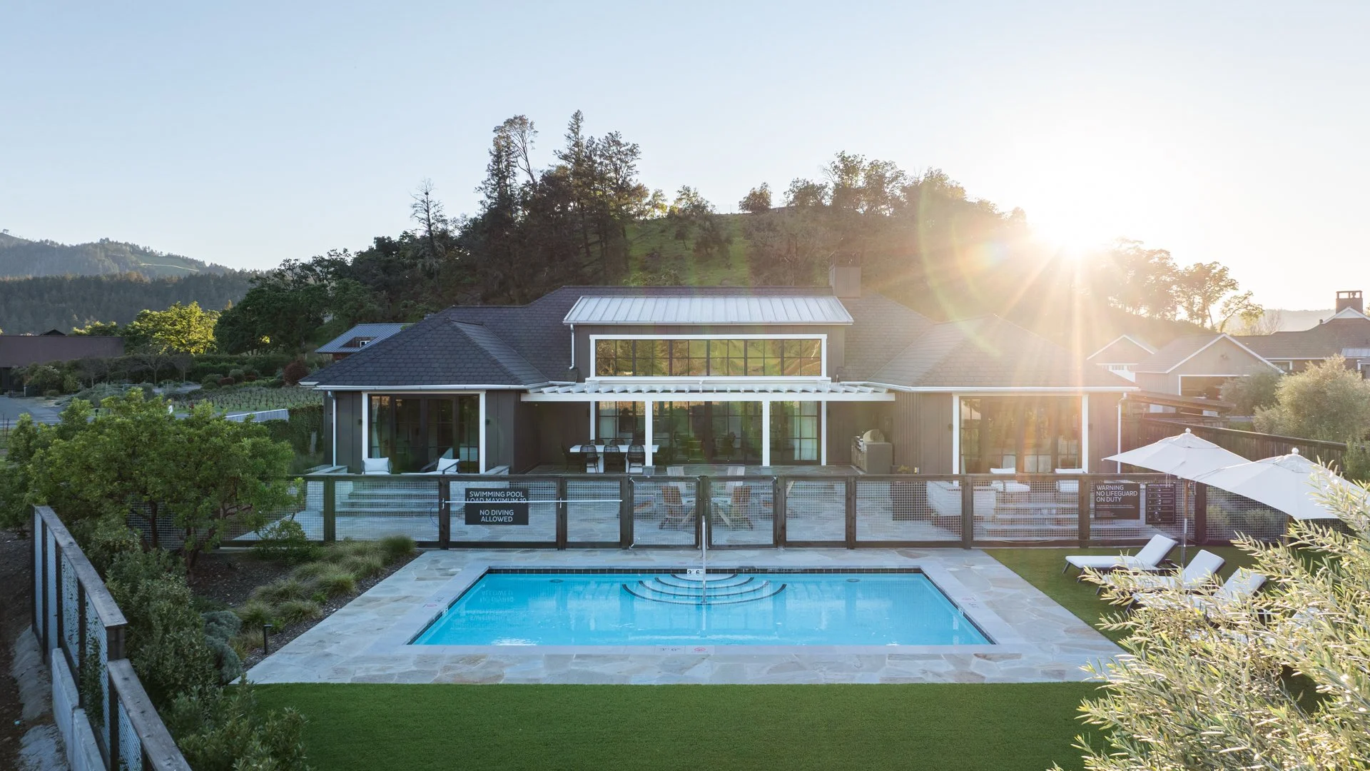 A backyard with a swimming pool in front of a modern house, surrounded by a black fence and adjacent to trees and hillside, under a bright setting sun