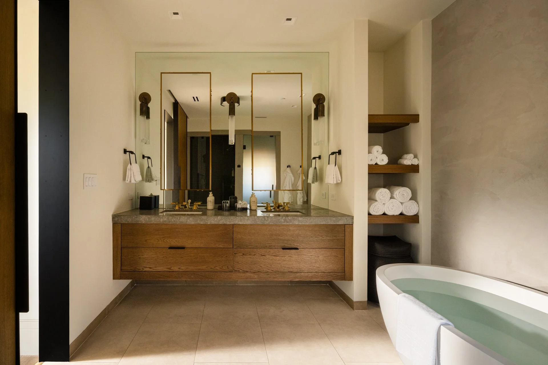 Modern bathroom with a large mirror above a double vanity with a marble countertop, open shelves filled with rolled white towels, and a freestanding bathtub with a towel hanging over its side.