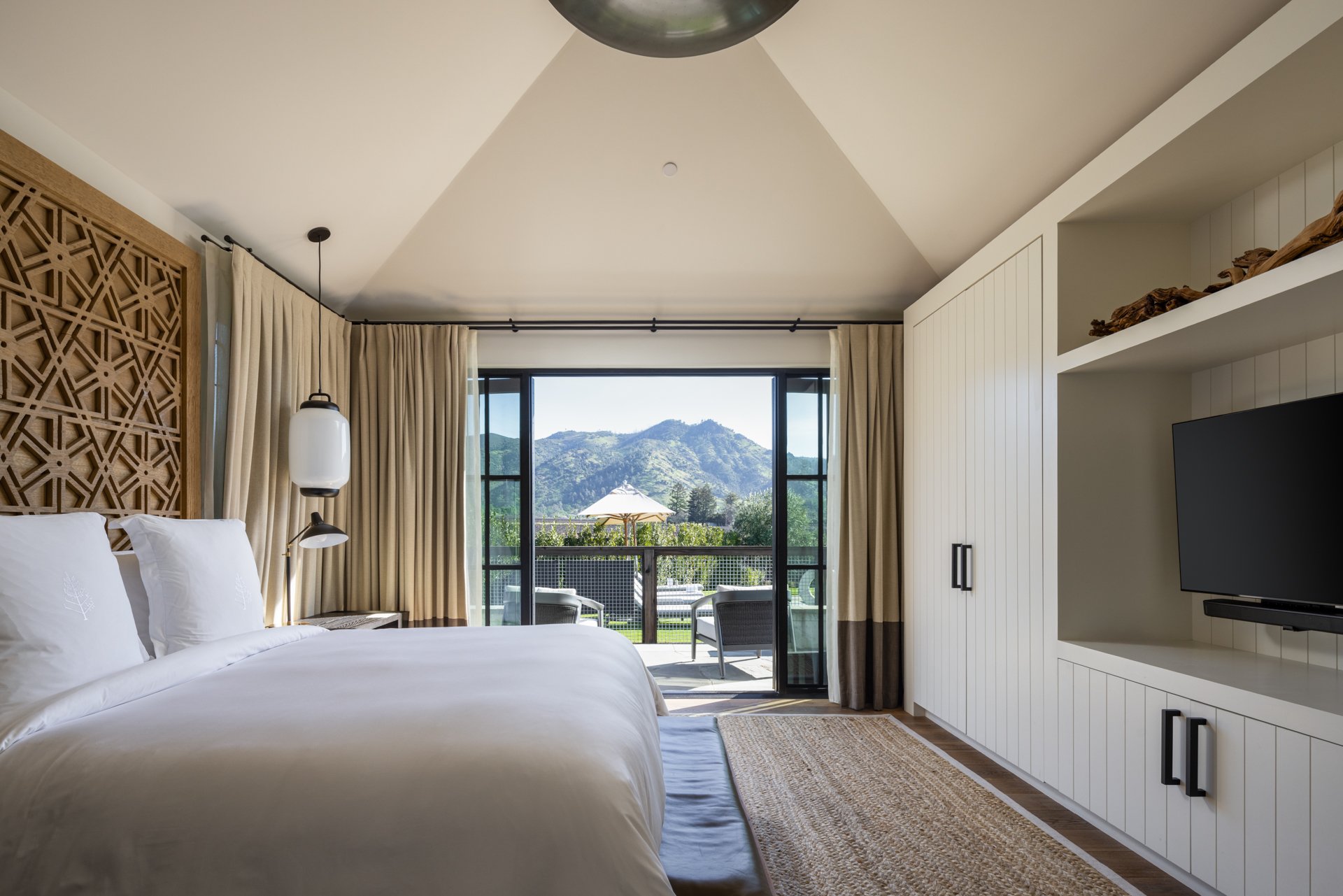 A bedroom with a large bed, intricate headboard, bedside lamp, balcony doors with curtains, outdoor patio with chairs and umbrella, mountain view, white built-in entertainment center, and a TV.