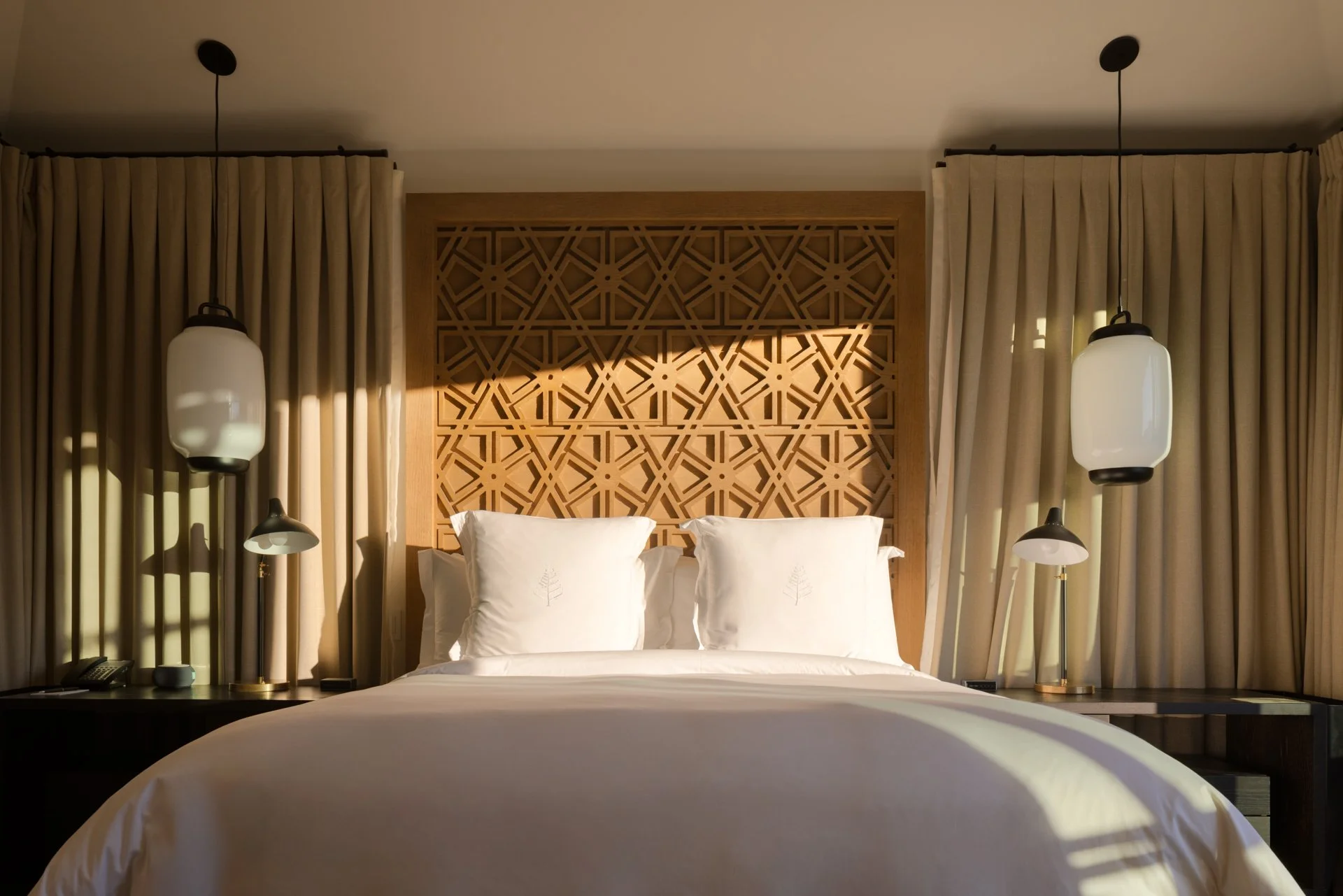 A hotel bedroom with a neatly made bed, white pillows, and an intricate wooden headboard. Two pendant lights hang on either side of the bed, with curtains framing the window.