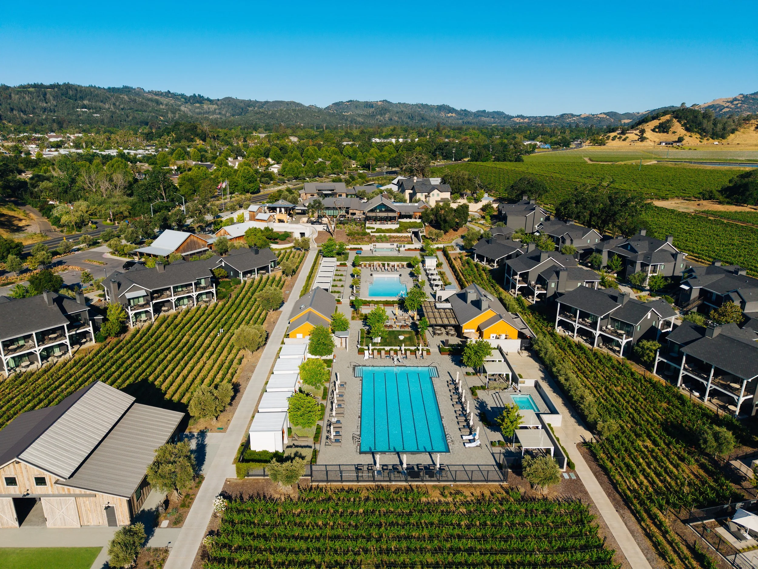 Aerial view of a community with swimming pools, surrounding green vineyards, residential buildings, and rolling hills in the background.