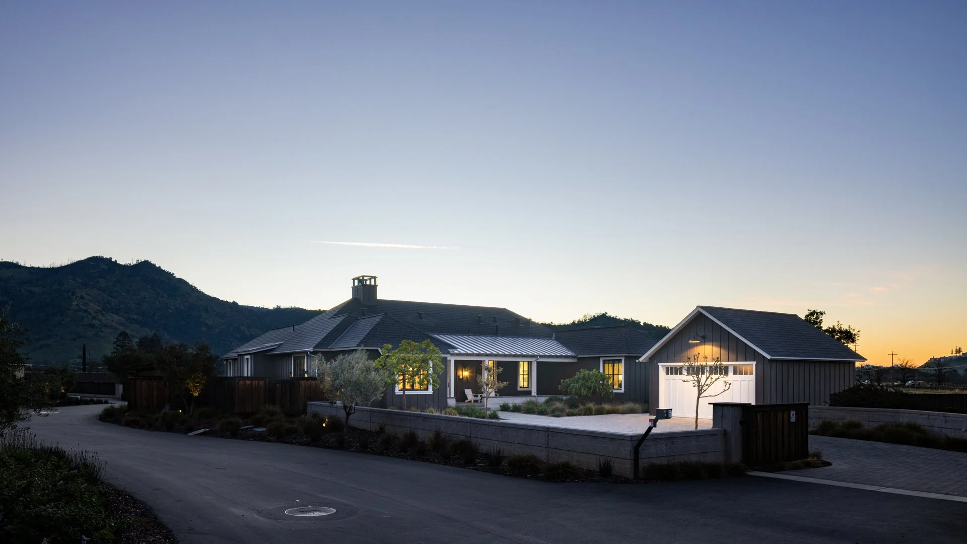 Modern house with lit windows and a garage, surrounded by trees and a driveway, during twilight with mountains in the background.
