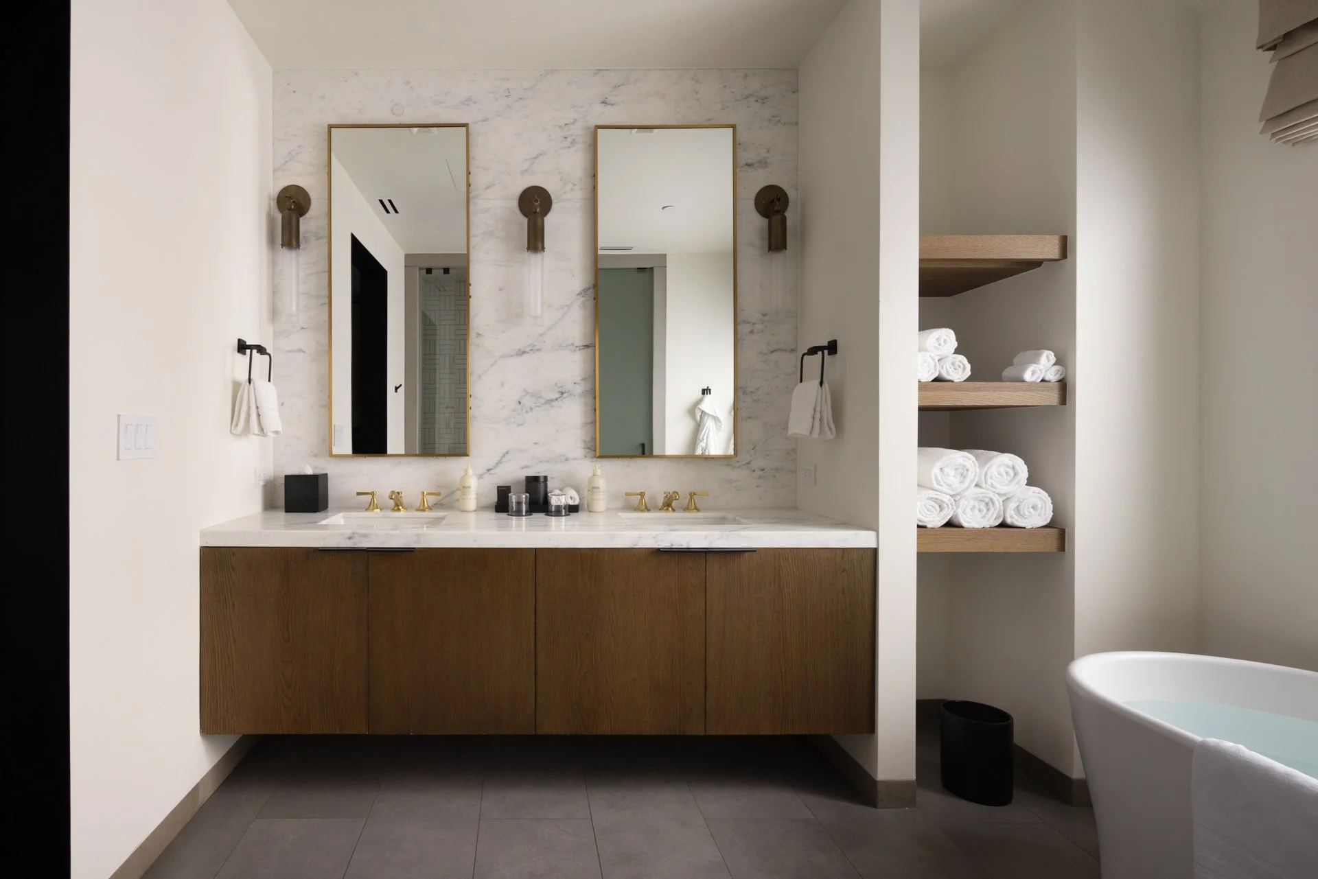 Modern bathroom with a double vanity, two mirrors, wall-mounted lights, towels, and shelves with rolled towels, and a bathtub.