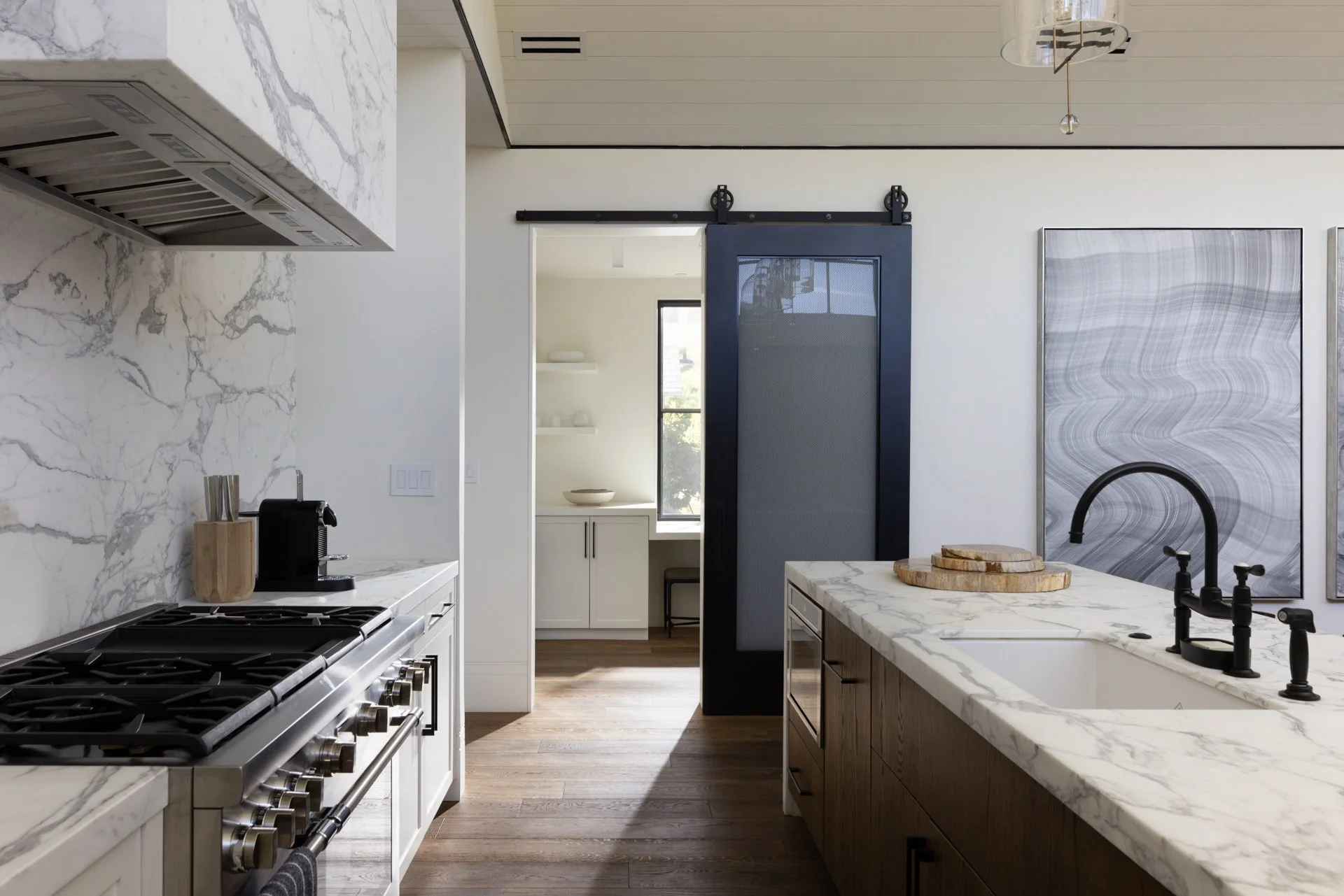 Modern kitchen with white marble countertops, a stainless steel stove, a black coffee machine, and a black sliding barn door. The kitchen includes white and wood cabinets, a sink with black faucet, and decorative art on the wall. There is a narrow ha