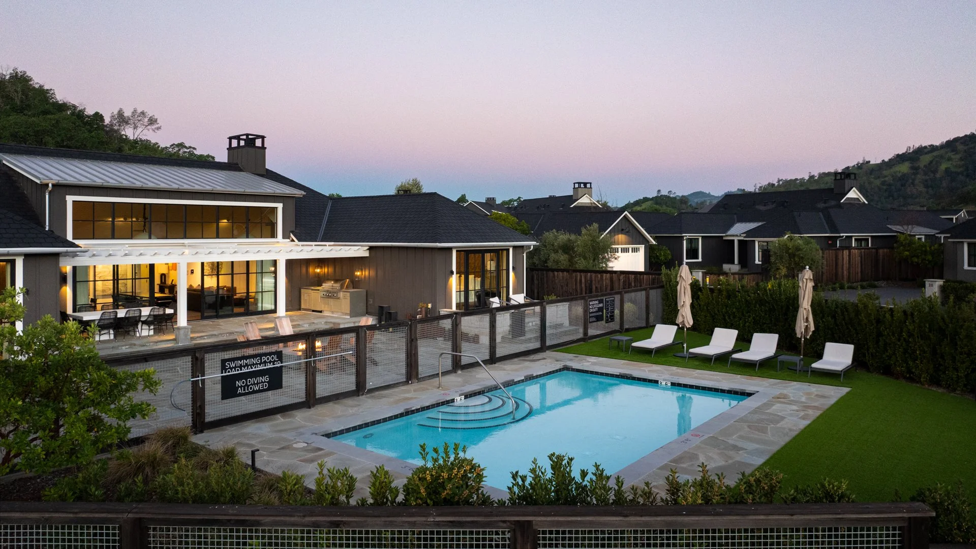 Relaxing backyard with a swimming pool, poolside lounge chairs with umbrellas, and a modern house with large windows, outdoor kitchen, and mountain landscape in the background at dusk.