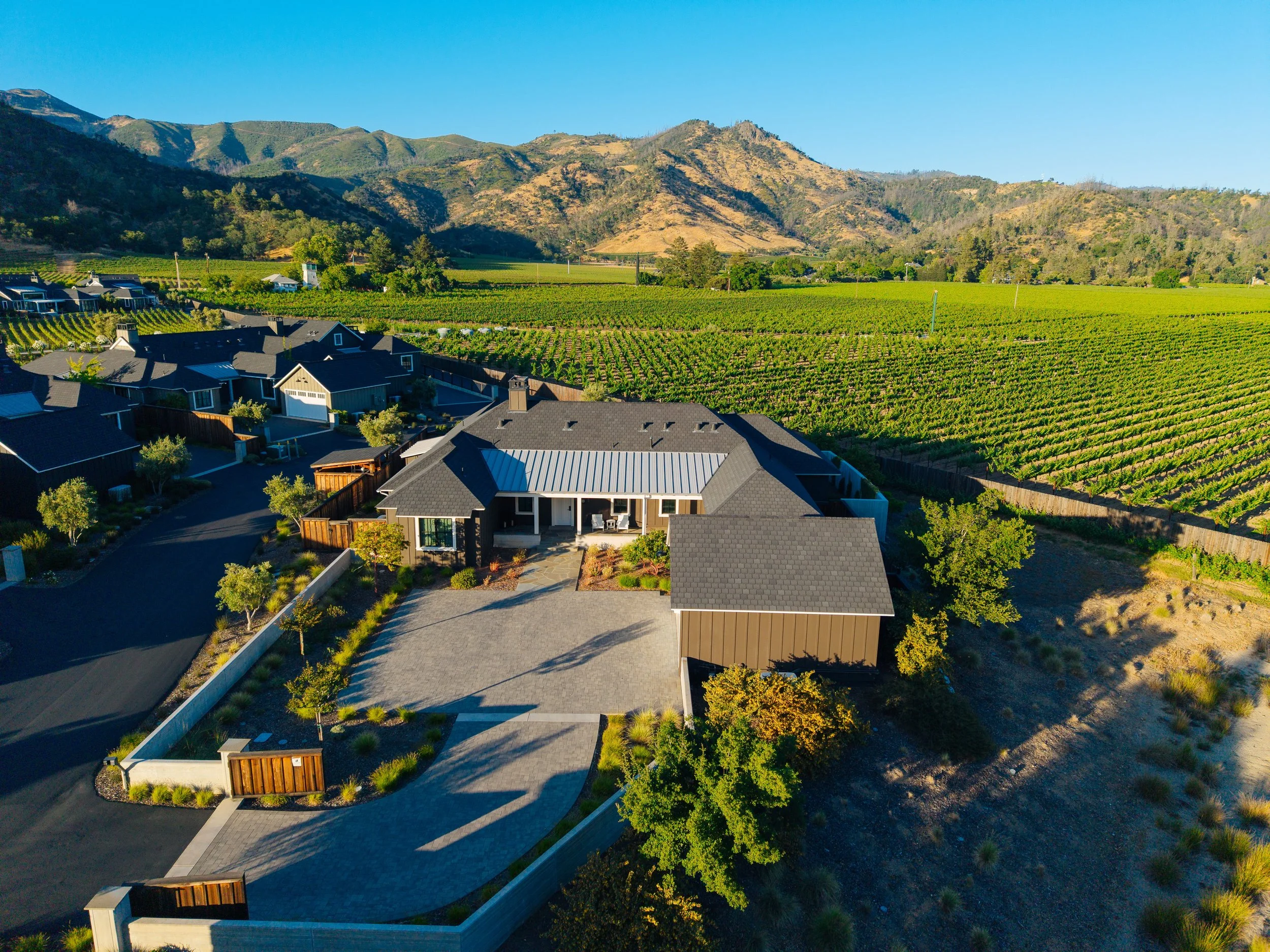 Aerial view of a modern house with a driveway and landscaped yard, surrounded by a suburban neighborhood with similar houses and expansive vineyards or farmland across the landscape, with mountains in the background.