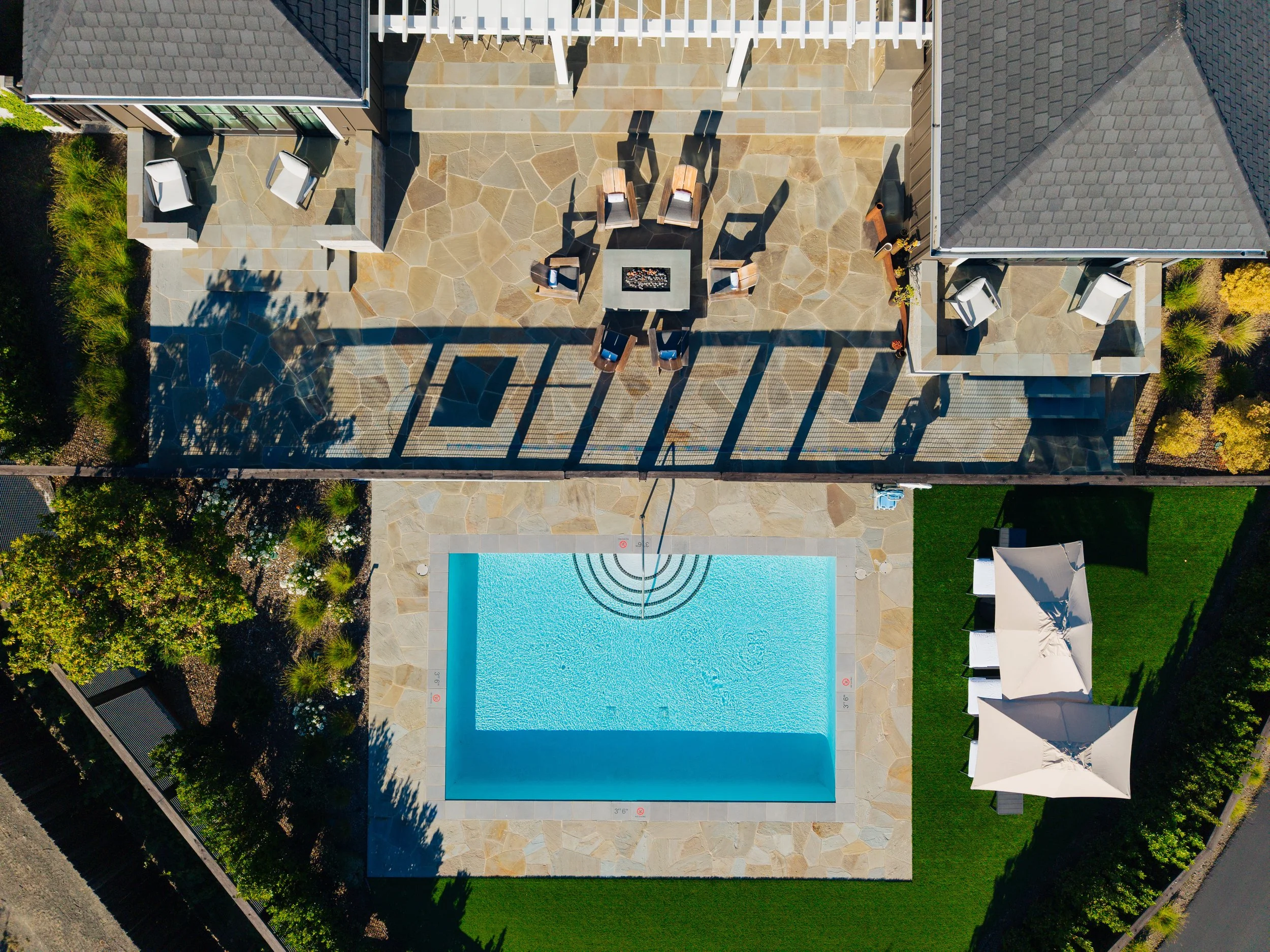 An aerial view of a backyard with a swimming pool, patio seating area with chairs and tables, and a gazebo on green grass.