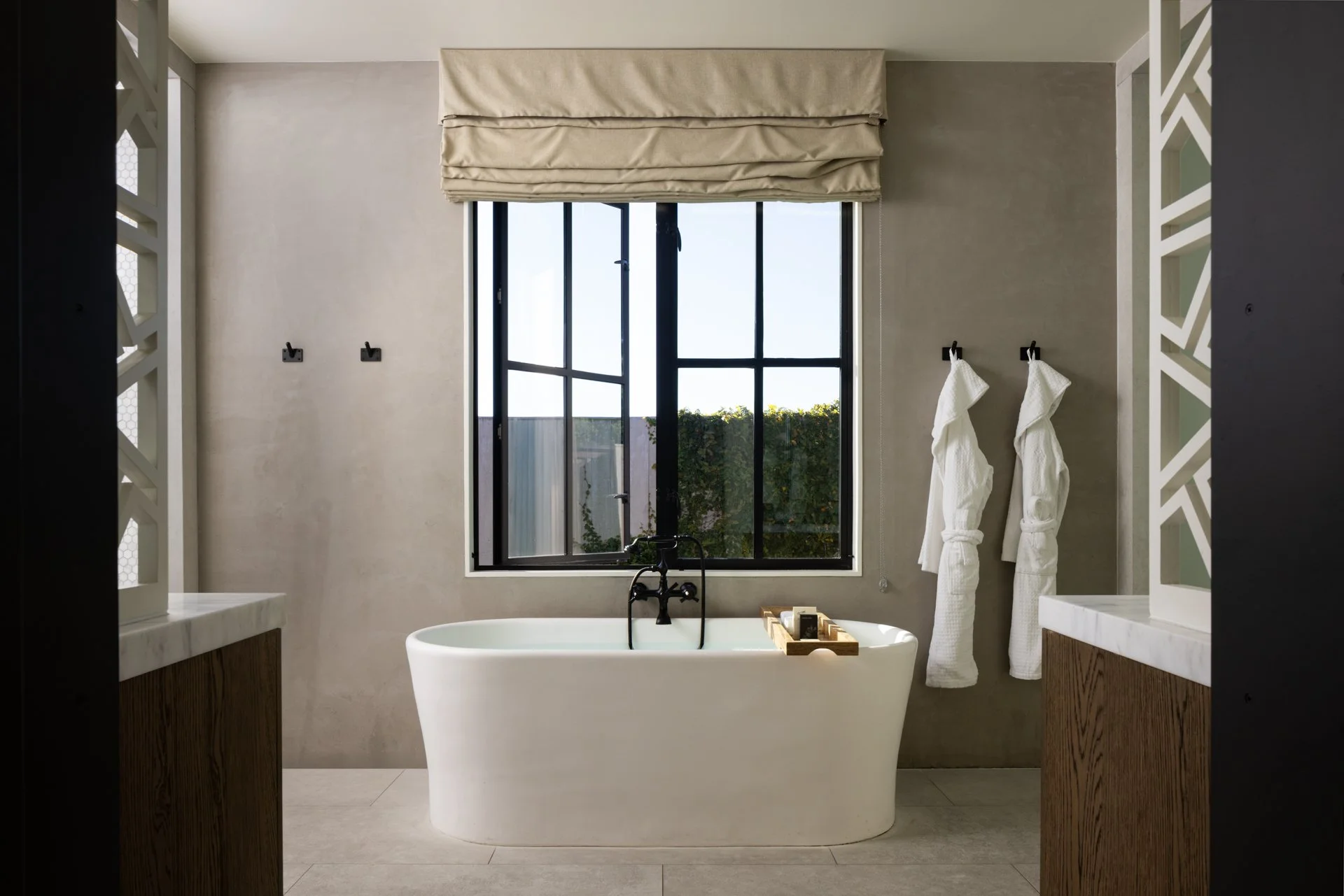 Modern bathroom with freestanding bathtub facing a window, two white bath robes hanging on hooks, and a tray with toiletries on the tub.