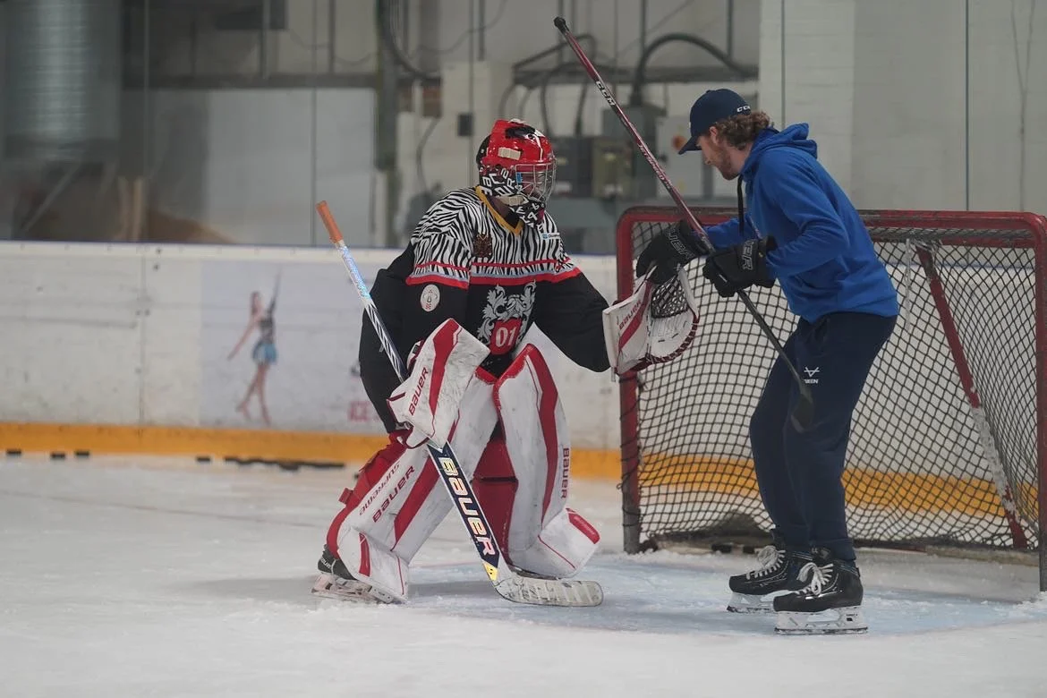 Hockey goalie in protective gear standing on ice rink near young man in blue hoodie leaning on hockey net.
