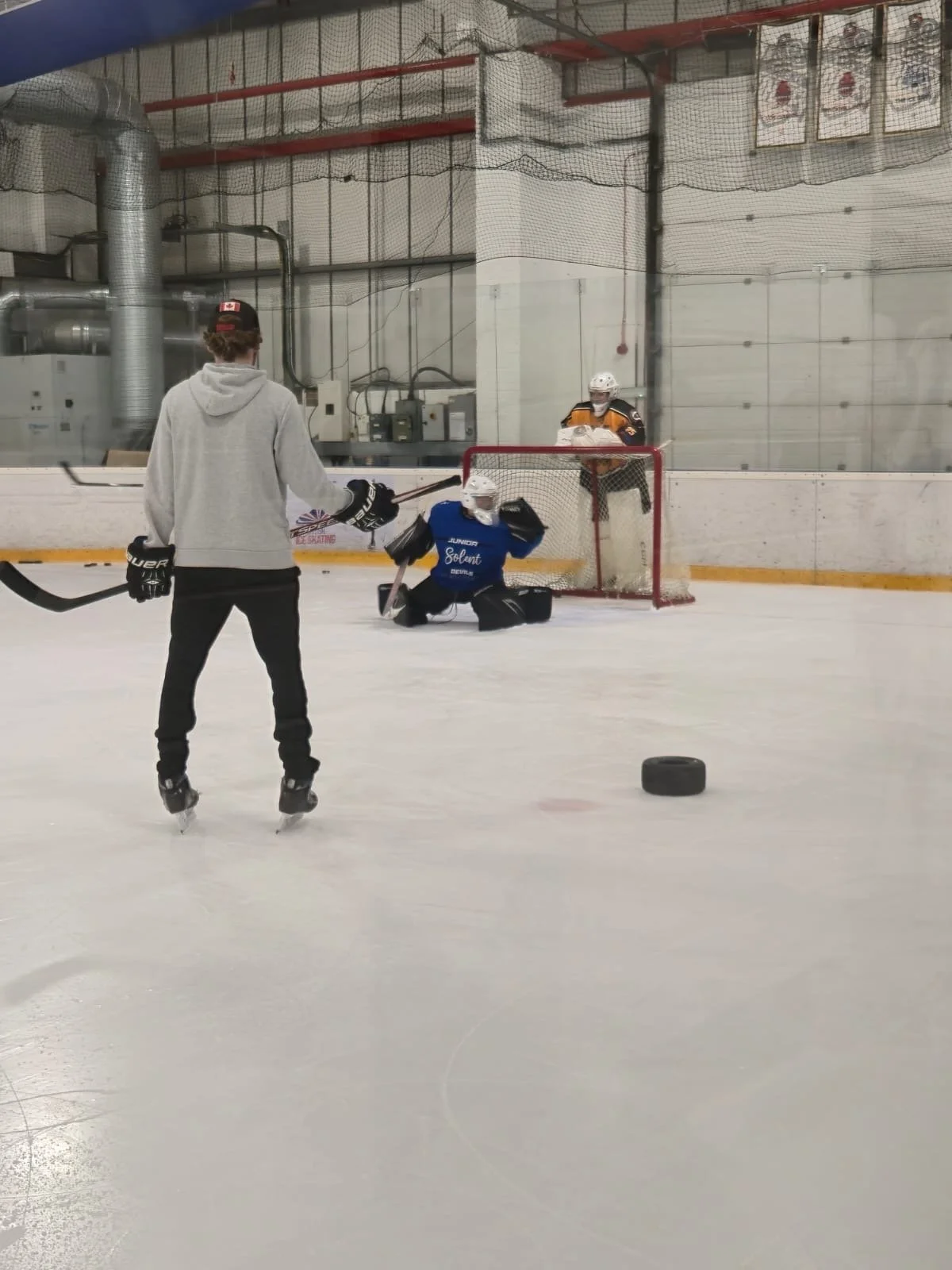 Hockey players practicing on an indoor ice rink. A skater in gray and black stands near a goalie who is kneeling with a glove extended, and a puck on the ice. Another player in an orange and black jersey stands behind the net.