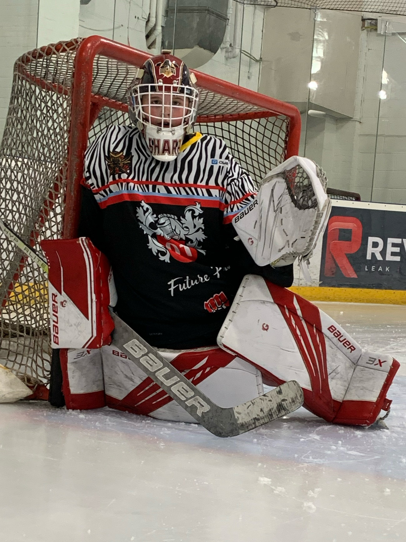 Ice hockey goalie in black and white jersey with red accents, standing in front of the goal on ice rink, wearing goalie pads, helmet, and gloves, holding a hockey stick.