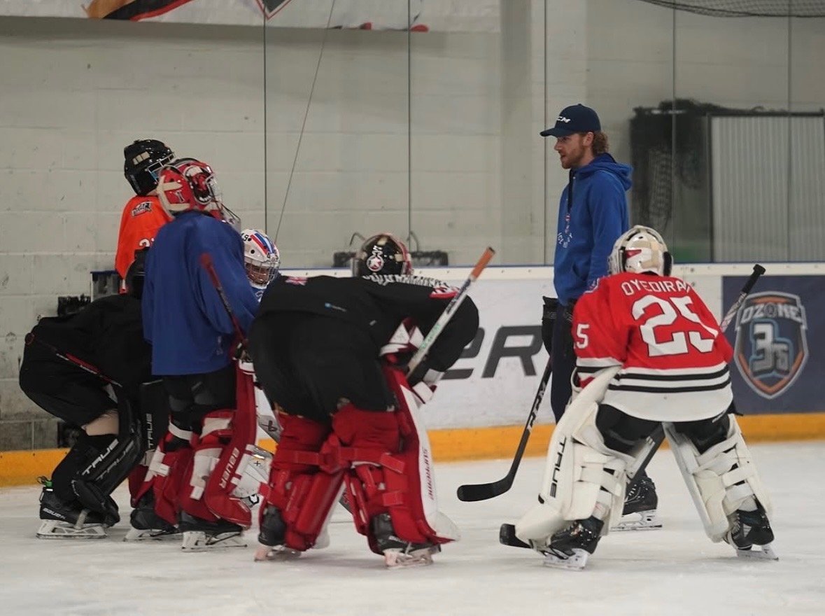 Hockey coach giving instructions to young hockey players on the ice rink, with players in goalie gear and helmets listening attentively.