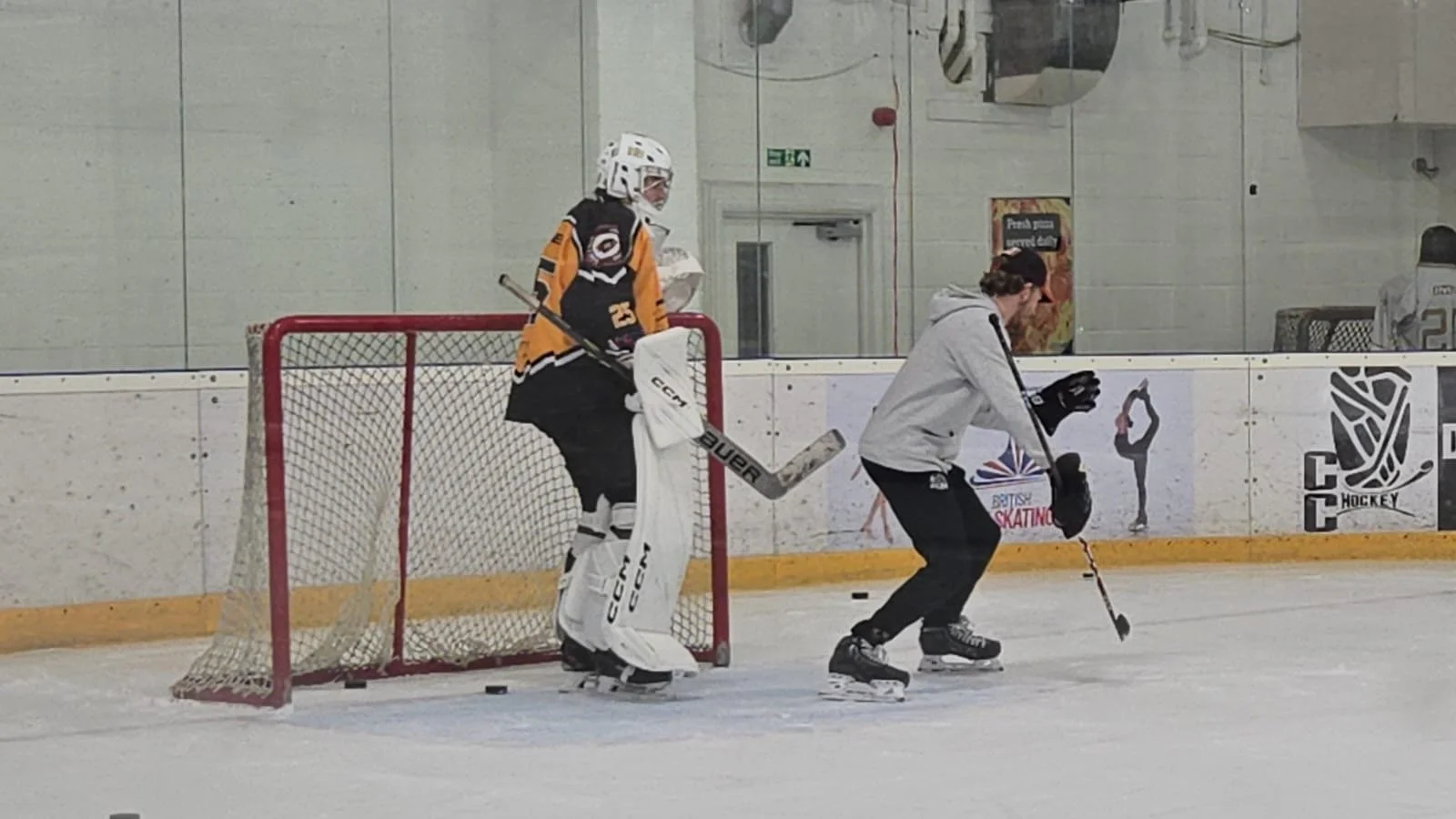 A hockey goalie in a yellow and black uniform and a coach in a gray hoodie practicing on an ice rink.