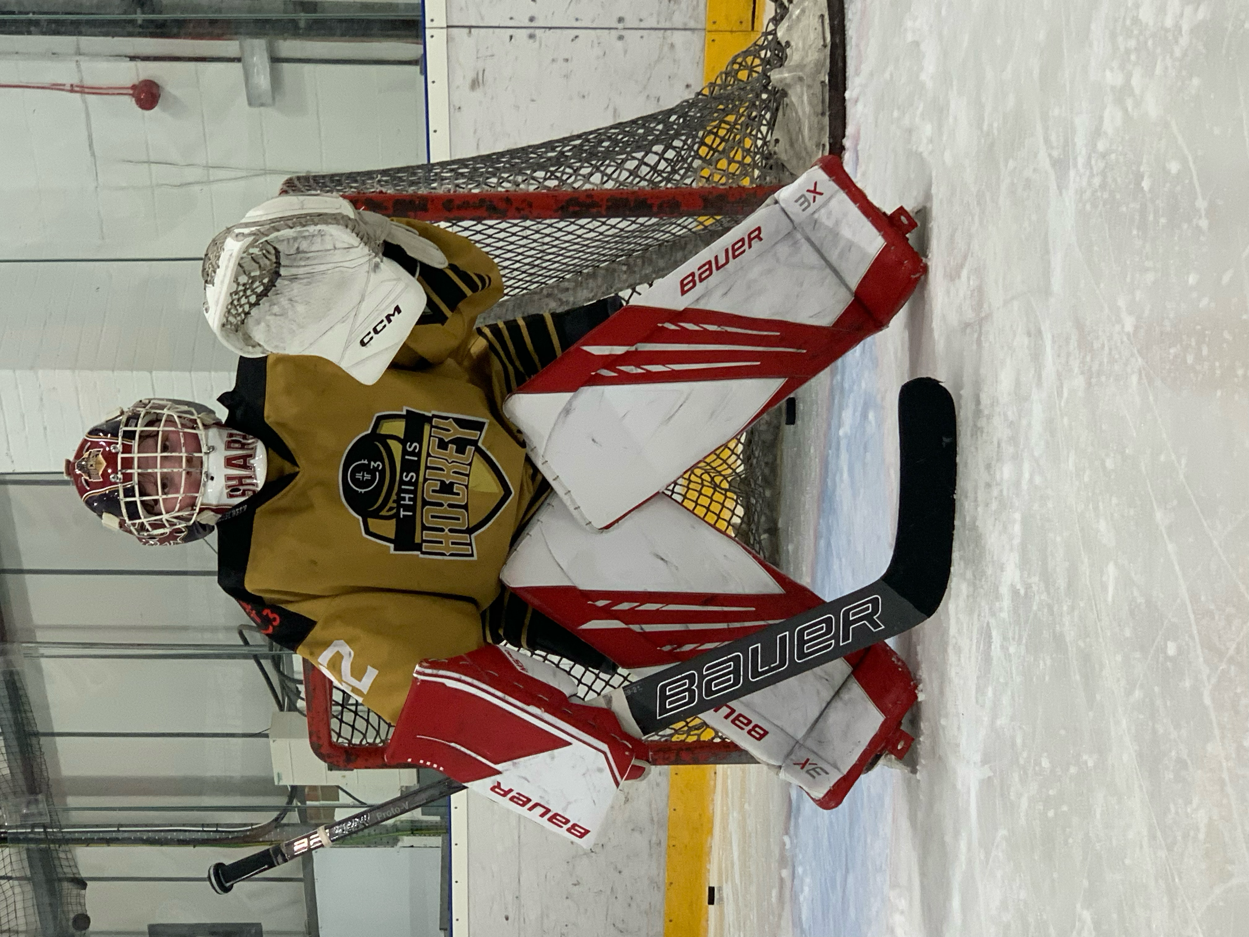Hockey goalie in a gold and black uniform sitting on the ice in front of the hockey net, wearing a white helmet and gloves, and holding a hockey stick.