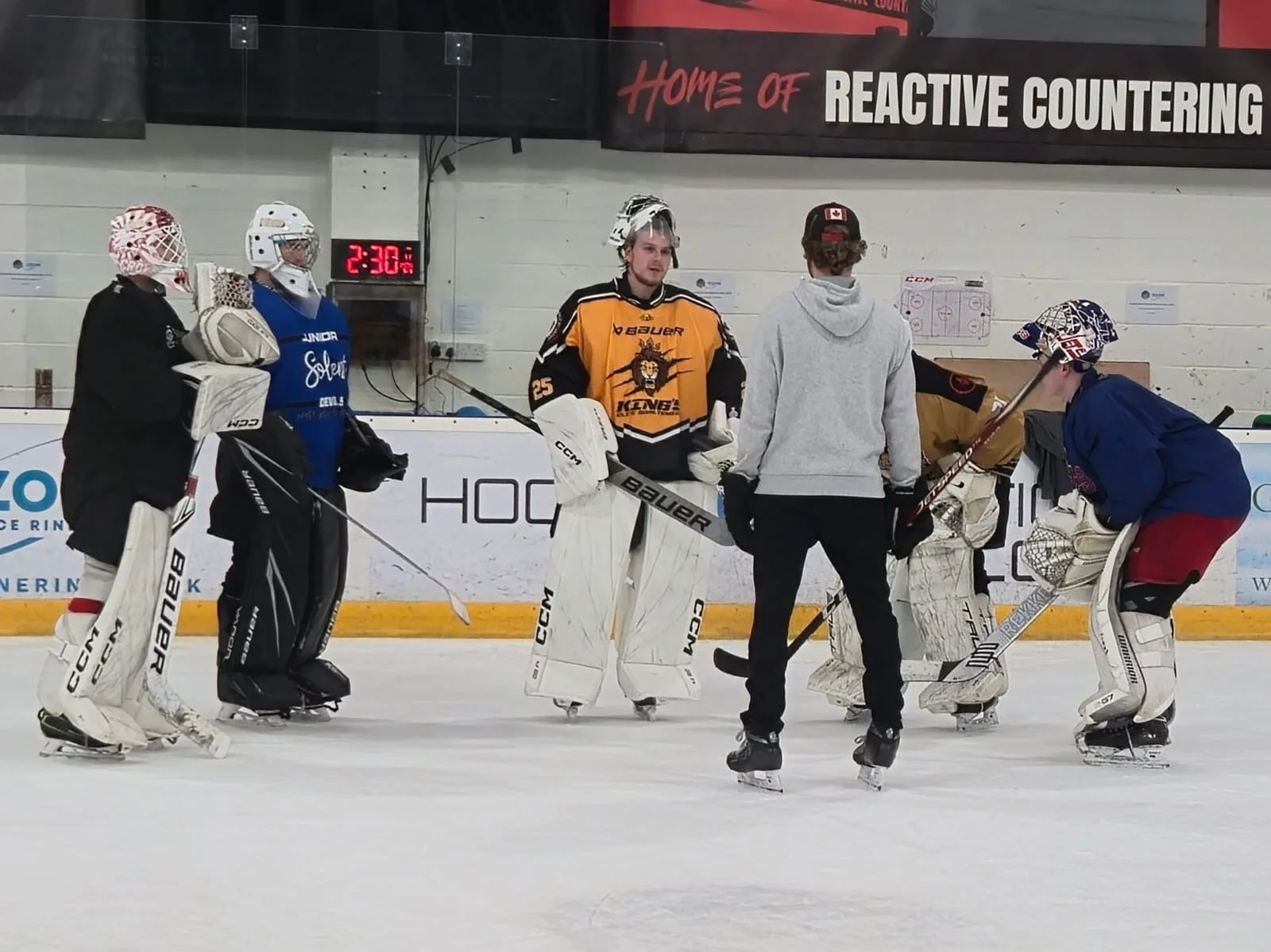 Hockey players and coaches on ice rink during practice or game, with goalie in blue jersey and referees in recent gear, in indoor hockey arena.